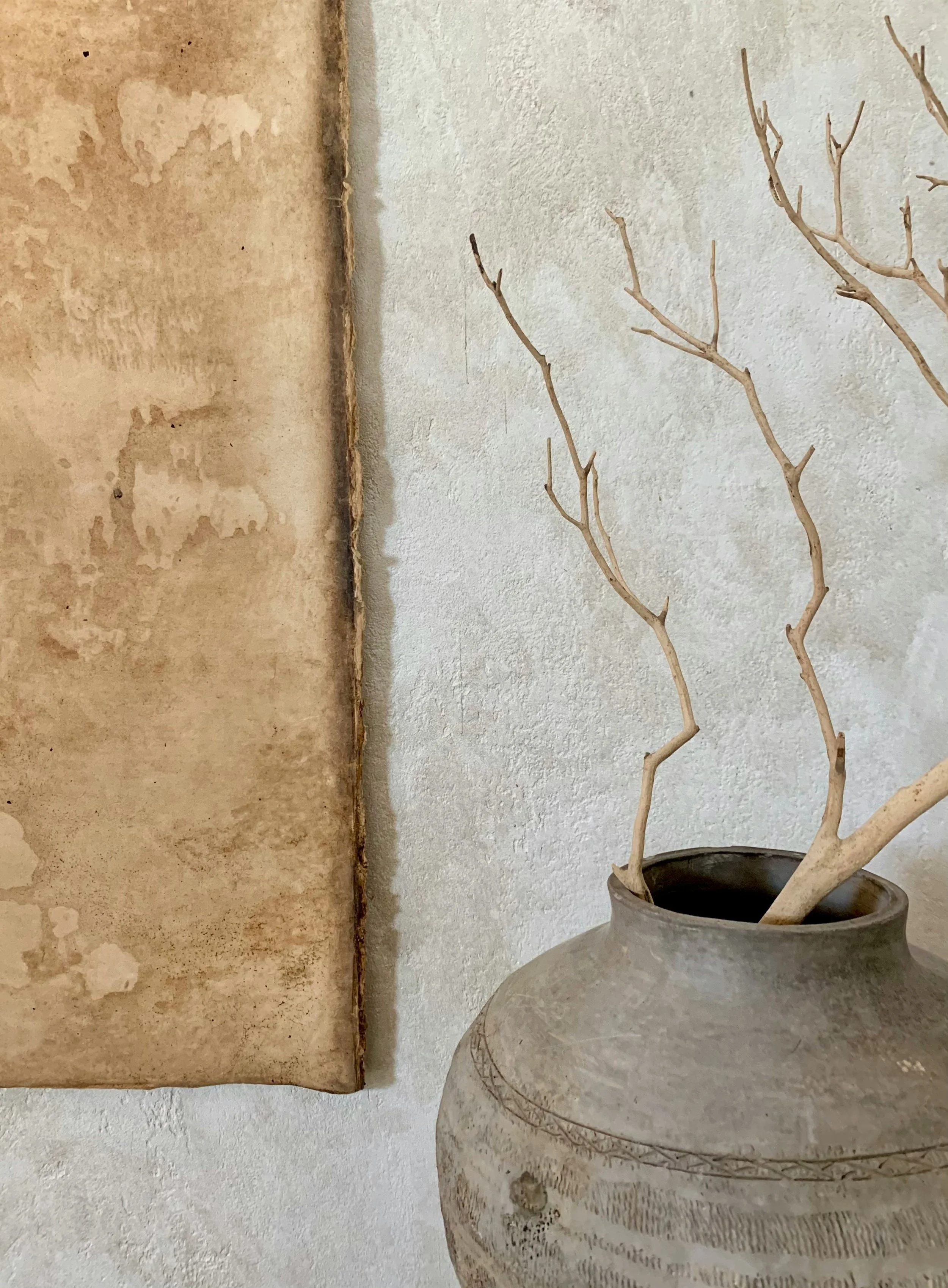 A large, round, gray ceramic vase with concentric circle and line patterns holds dry, leafless branches. The background consists of a rough, light-colored wall and a partially visible, old, weathered parchment or fabric hanging on the left side.
