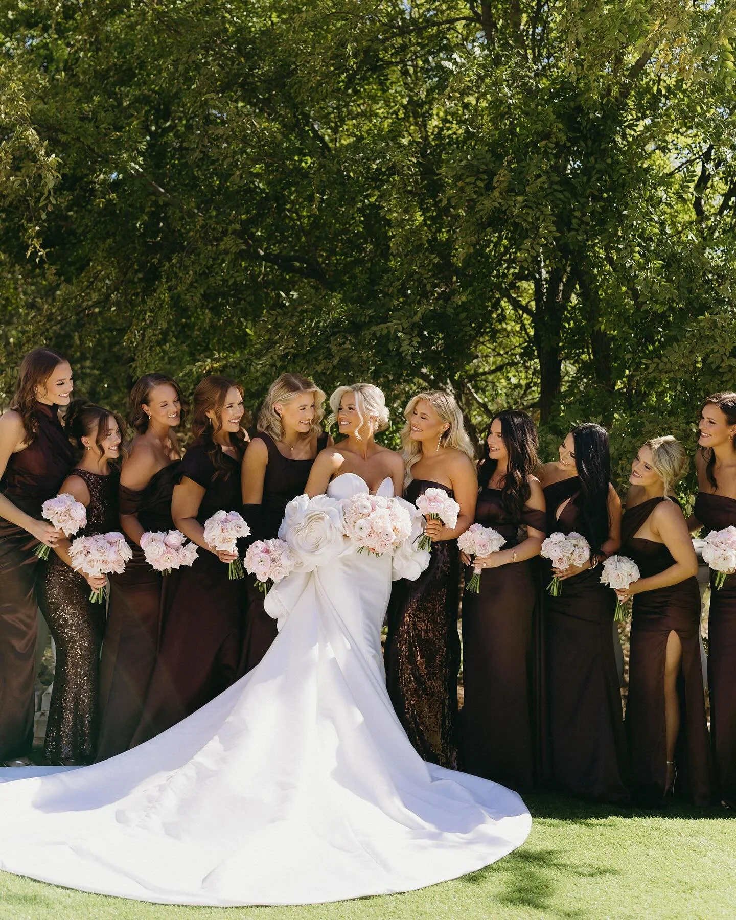 A bride in a white wedding gown stands surrounded by bridesmaids in black dresses holding bouquets, outdoors under a green leafy canopy.