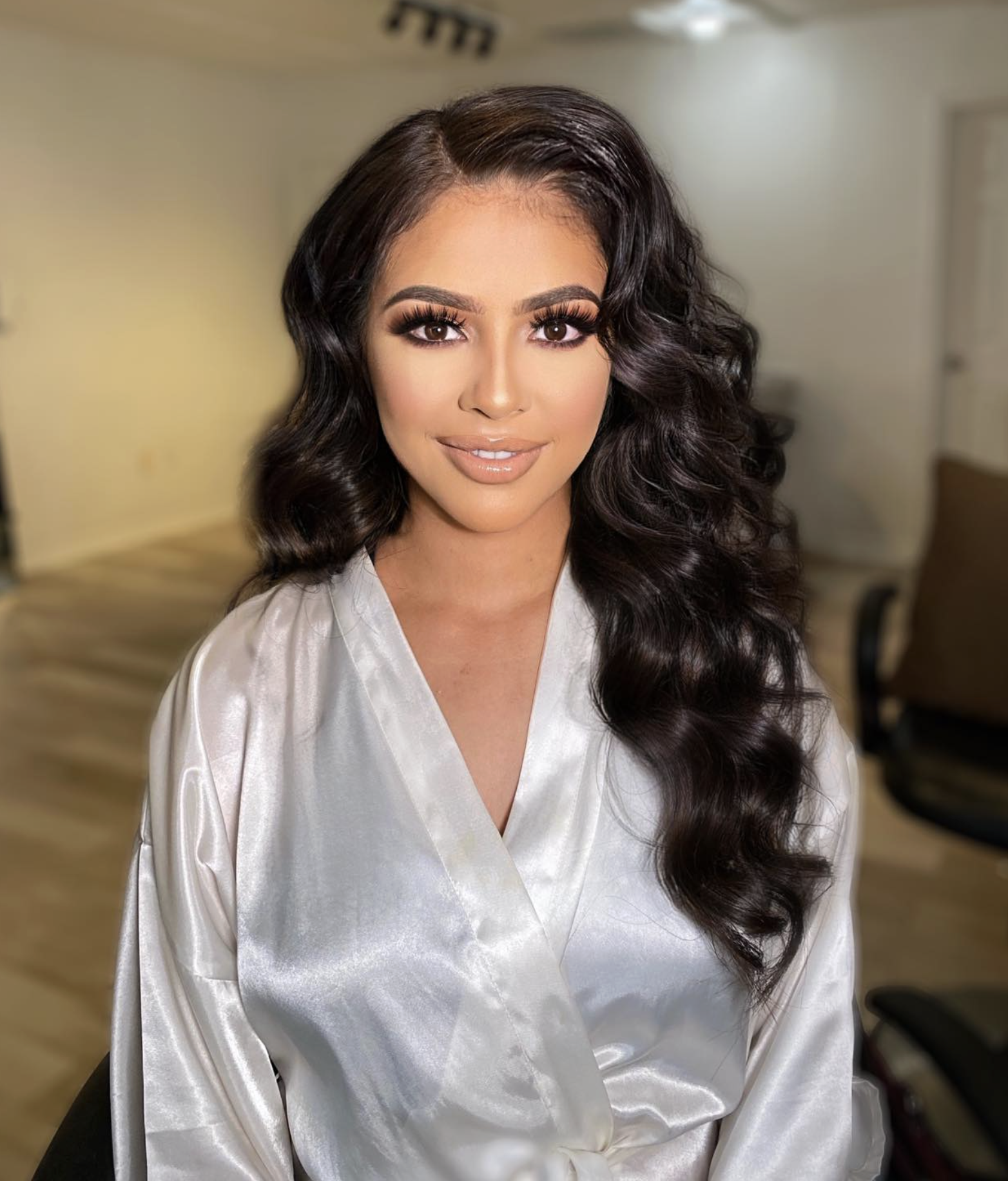 Close-up of a woman with long, dark wavy hair, wearing a white satin robe, in a room with a blurred background.