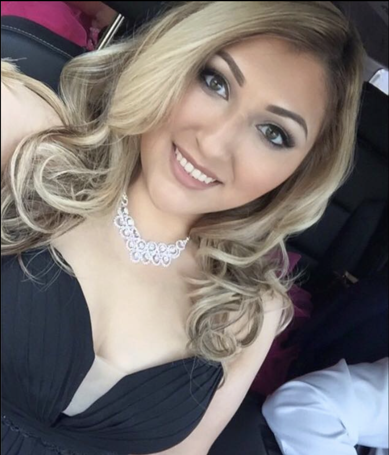A smiling woman with blonde curly hair wearing a black dress and a white lace necklace, sitting in a car.