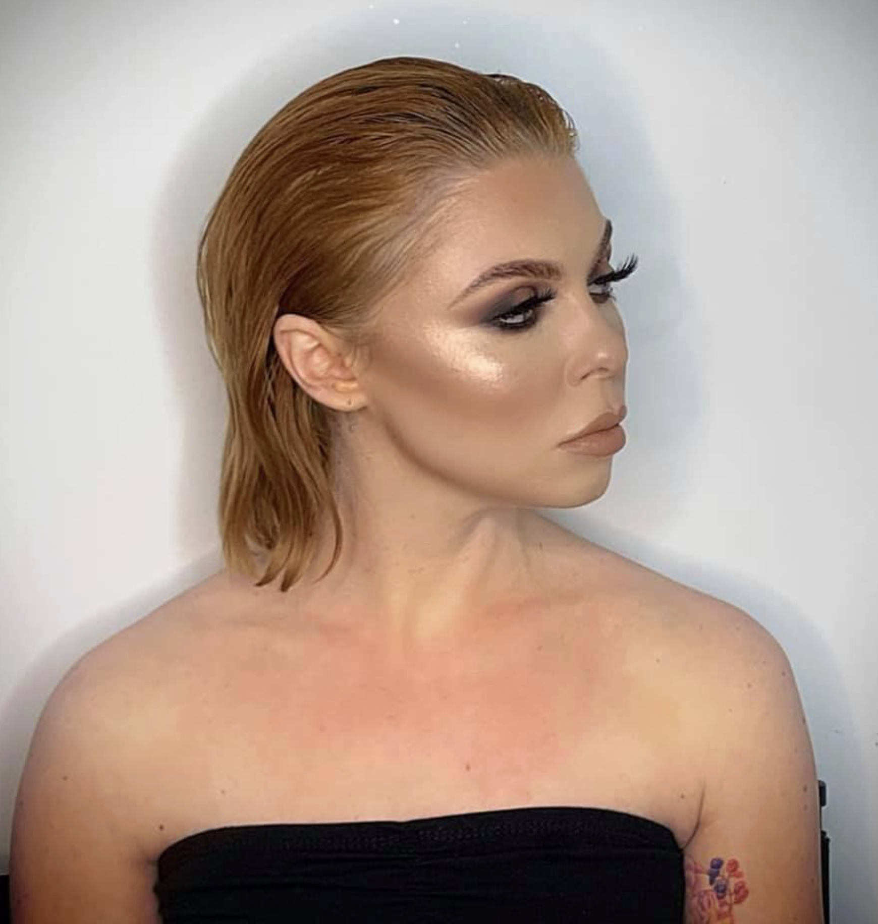 A woman with short, wet-looking, reddish-brown hair styled back, wearing makeup with dark, smoky eyeshadow, highlighter on her cheekbones, and nude lipstick, against a plain white background.