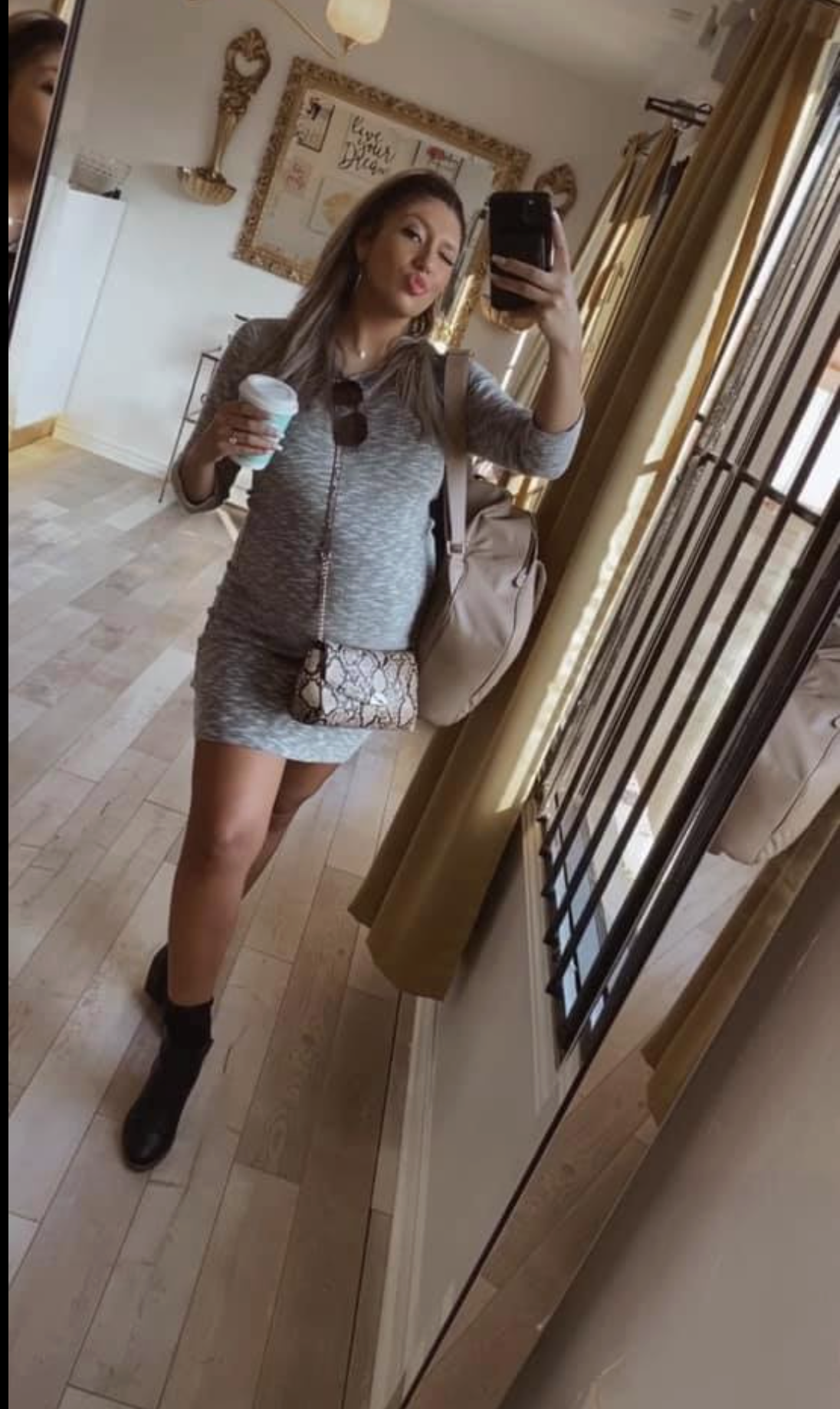 Woman with long hair wearing a grey zip-up top and matching shorts, holding a coffee cup, taking a mirror selfie in a room with wooden flooring and a large mirror.