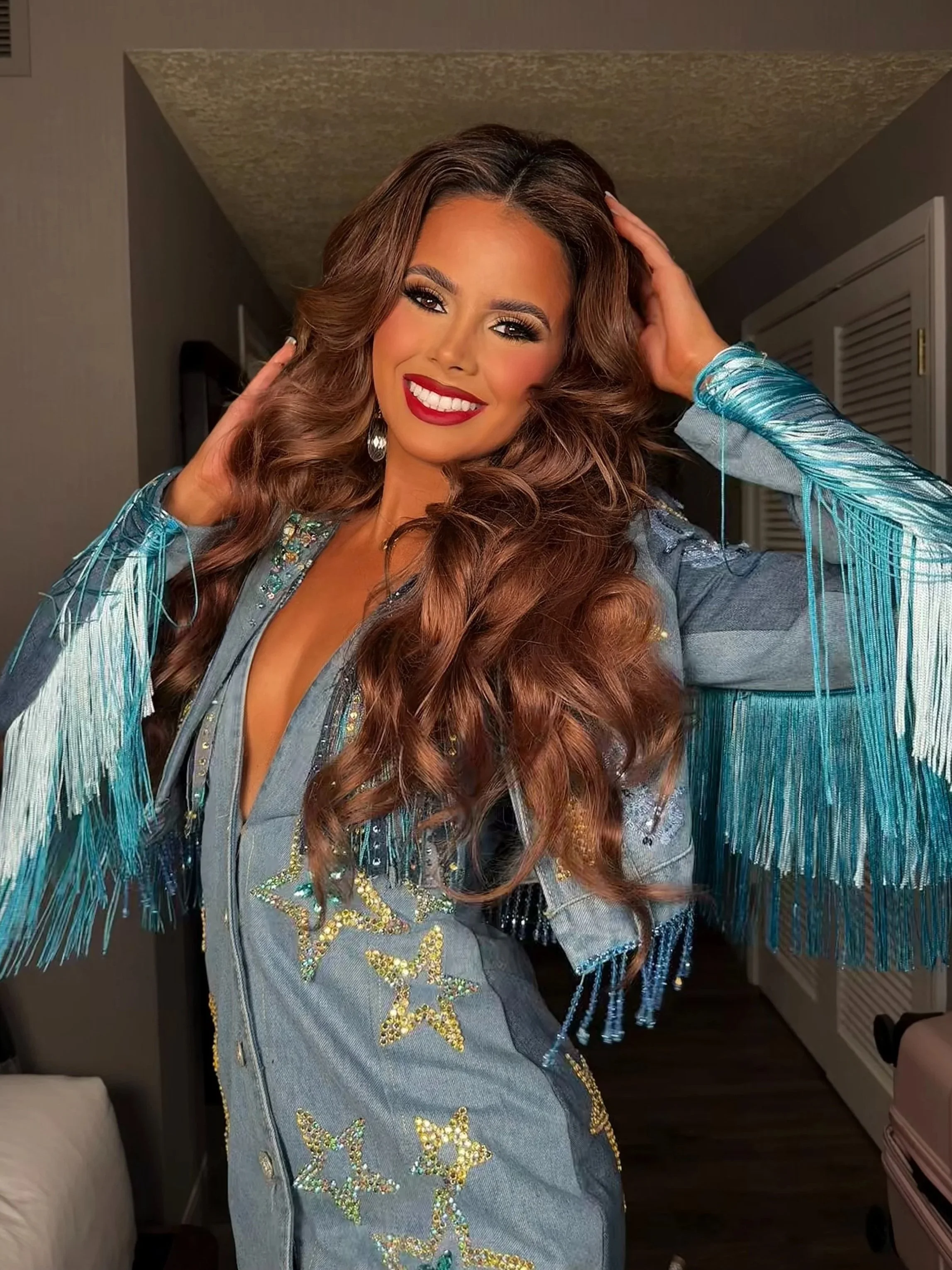 Smiling woman with long wavy brown hair in a star-patterned denim jacket with professional event makeup."