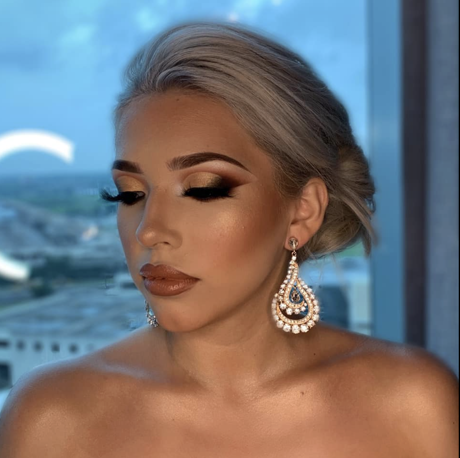 A woman with silver hair and makeup, wearing large, ornate earrings, appears to be in a high-rise building with a city view behind her.