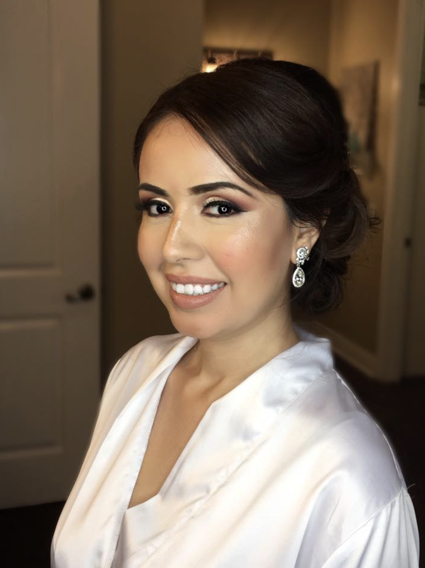 Close-up of a woman with dark hair styled in an updo, wearing makeup and earrings, smiling in an indoor setting.