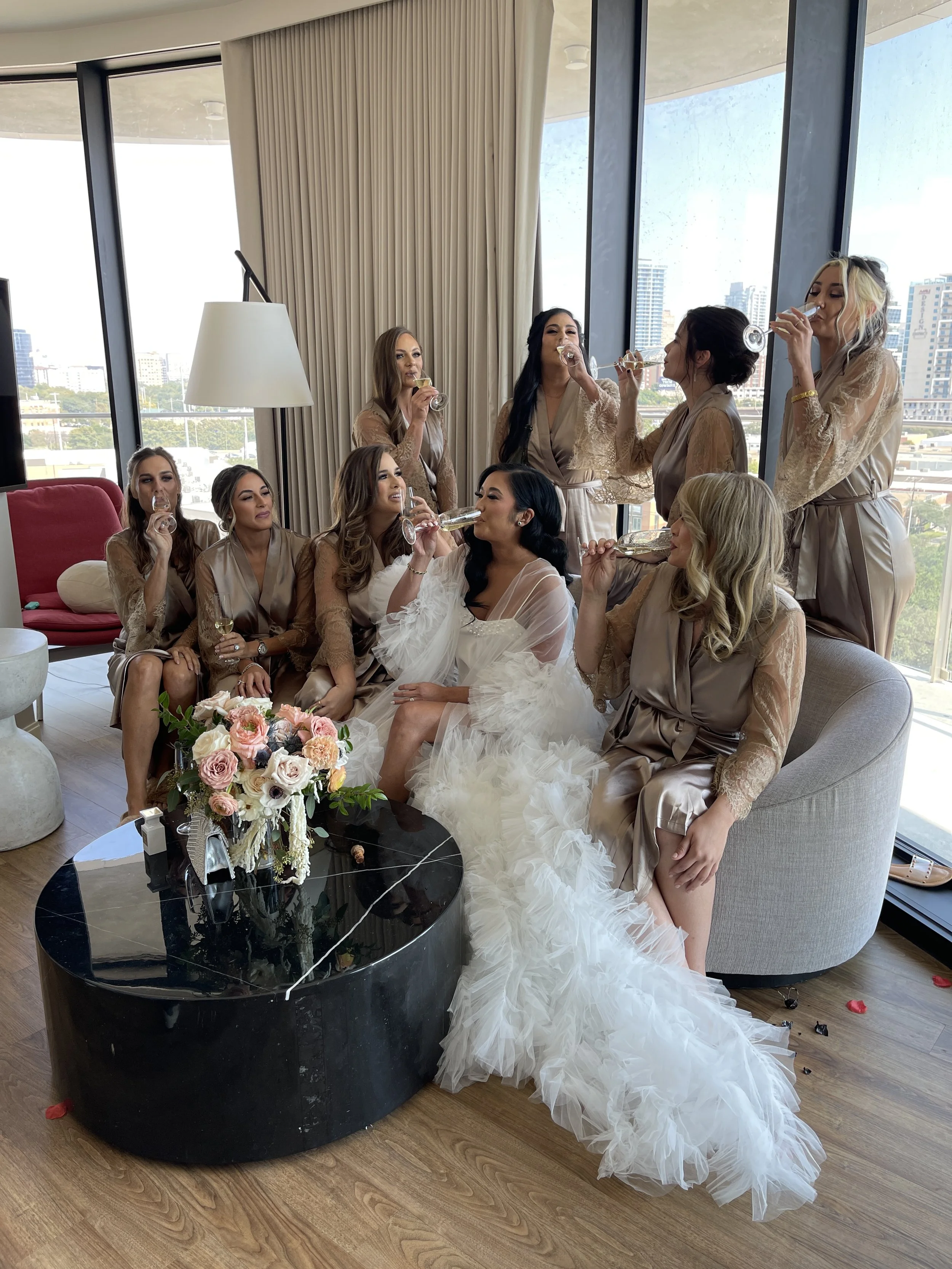 A bride and her bridesmaids toasting with champagne in a luxurious hotel room with floor-to-ceiling windows.