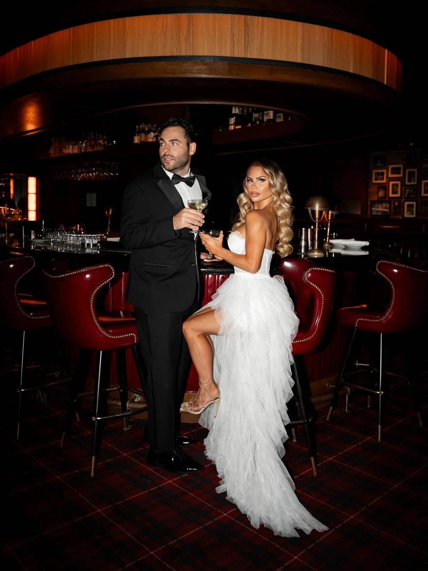 A man in a tuxedo and a woman in a wedding dress standing at a bar, holding drinks in a dimly lit upscale bar or lounge.