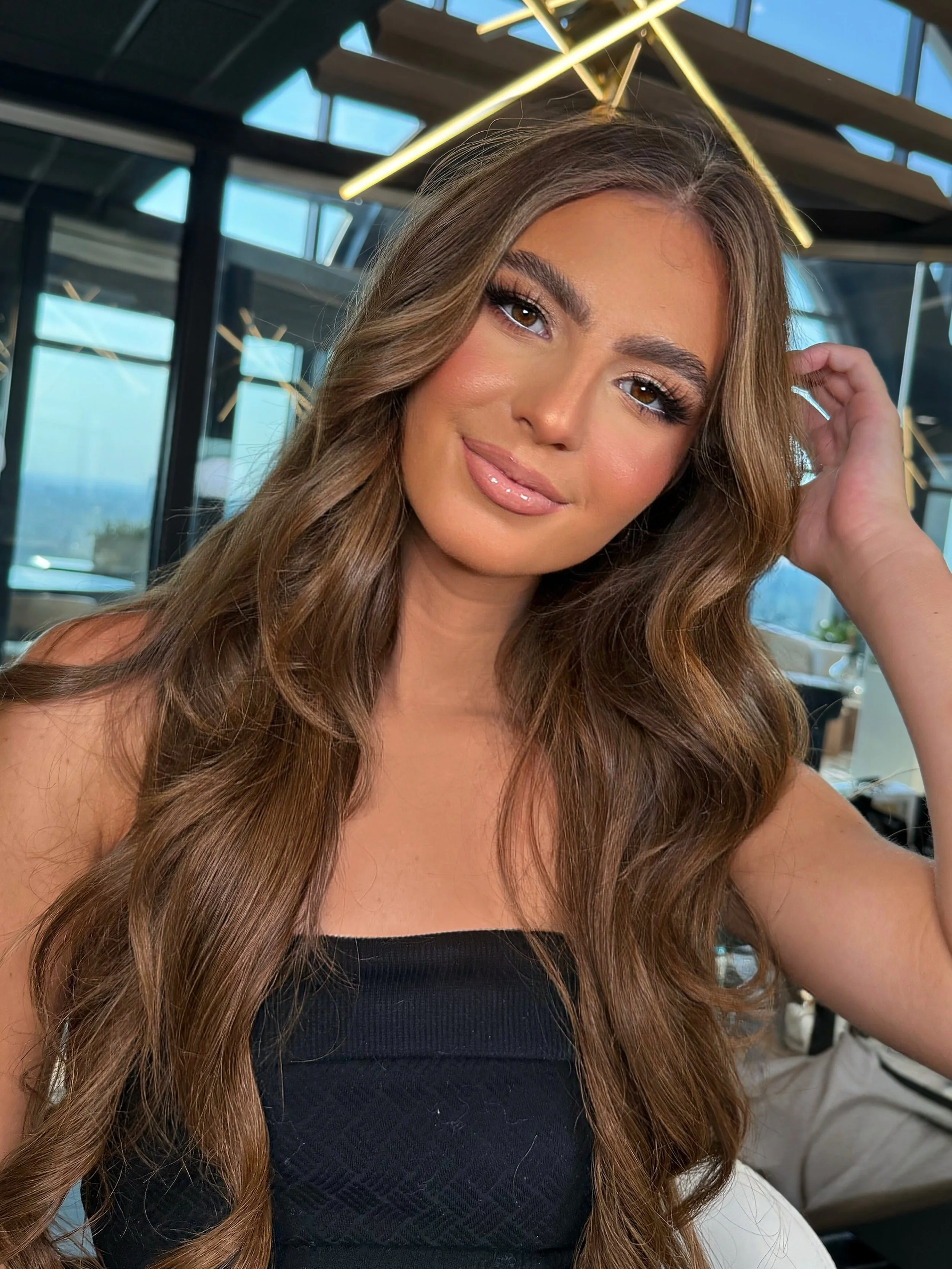 A young woman with long, wavy brown hair taking a selfie inside a modern building with large windows and geometric lighting fixtures overhead.