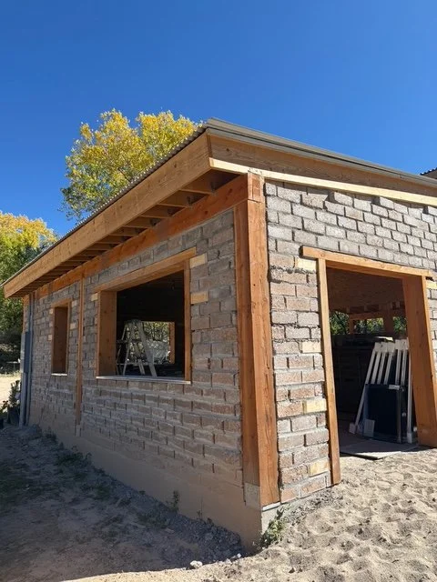 Construction site of a stabilized compressed earth block building with wooden framing, under blue sky with trees in the background.