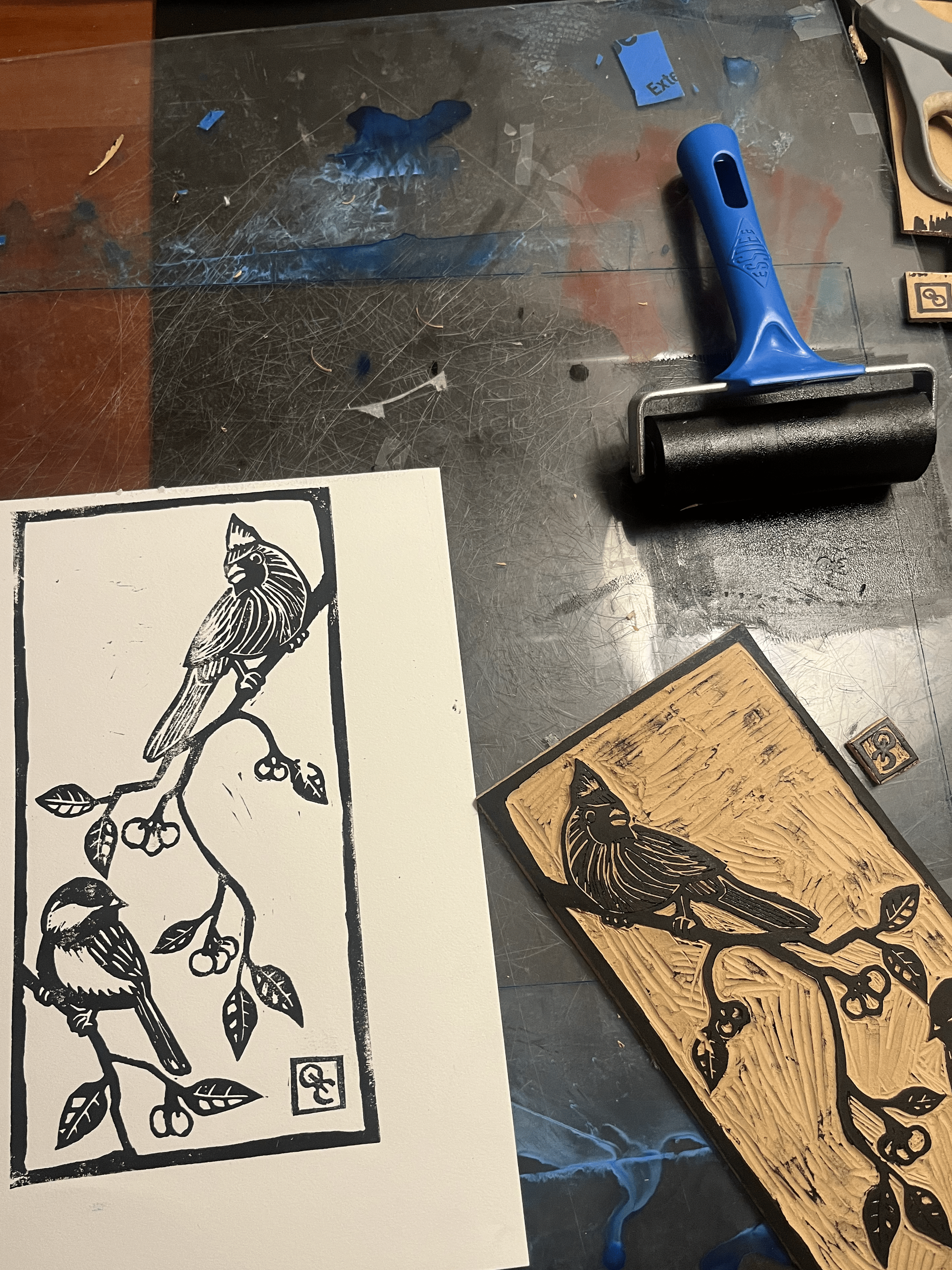 A rubber stamp of a bird sitting on a branch with berries, placed on a wooden block. The stamp's design is also printed on a piece of paper next to it.