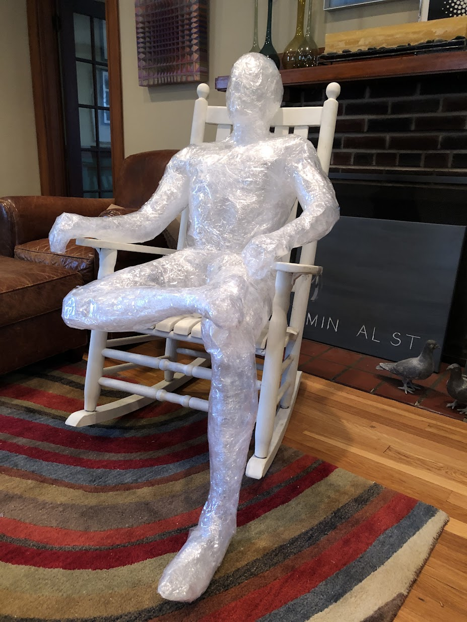 Sculpture of a human figure made from a transparent, crinkled material, seated in a rocking chair indoors.