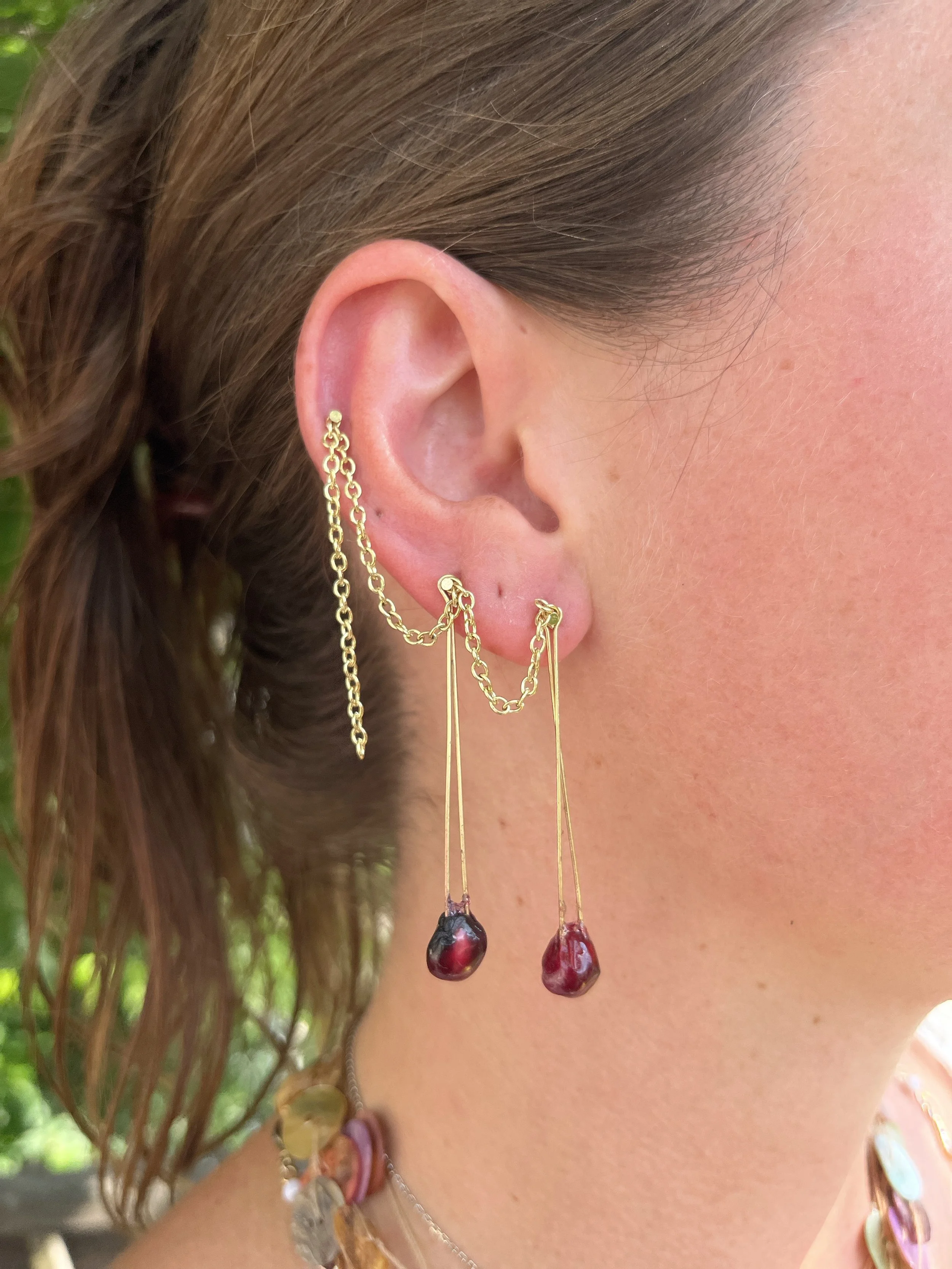 Close-up of a woman's ear wearing gold chain earrings with dark red beads and an additional piercing with a gold chain connecting two earrings.