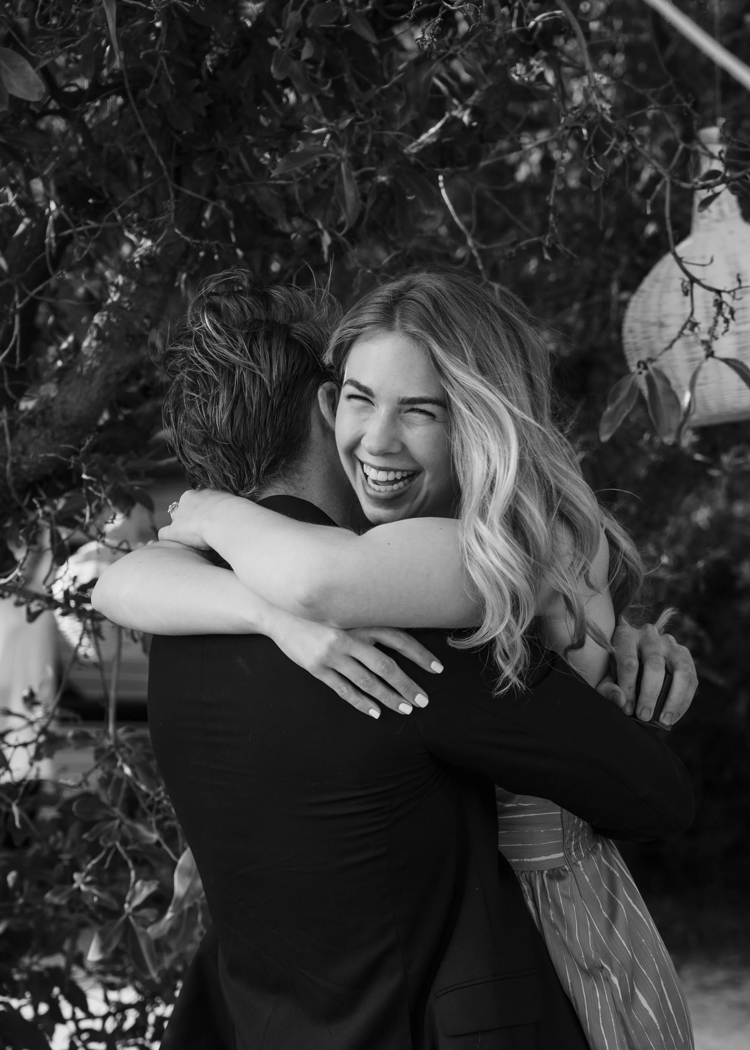 A joyful woman with wavy blonde hair hugging a man outdoors. She is smiling broadly and their faces are close together. The background features leafy foliage.