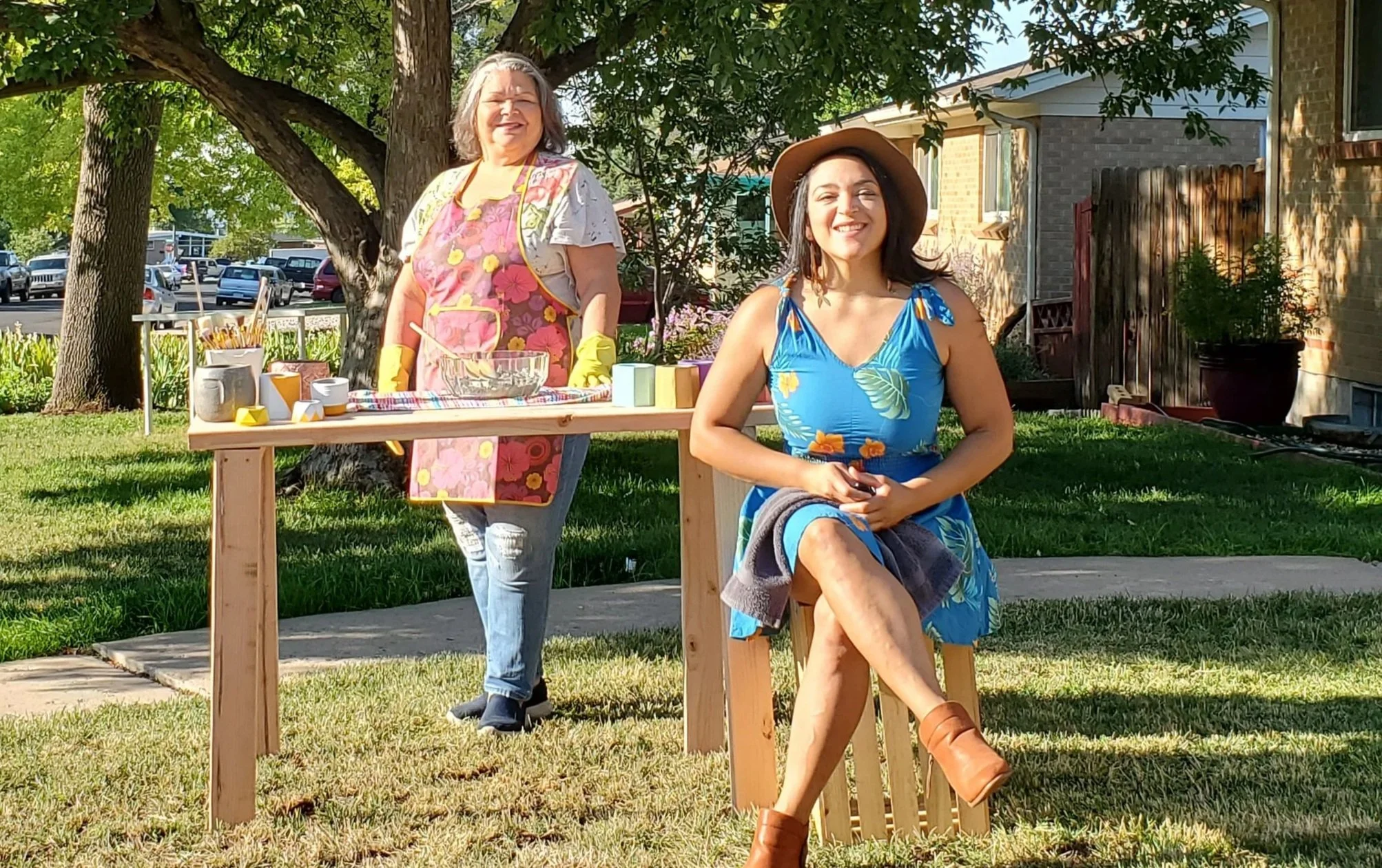 Two women in a backyard, one standing behind a table wearing an apron and gloves, the other sitting on a wooden chair in a blue floral dress and sun hat, smiling in sunlight with trees and houses in the background.