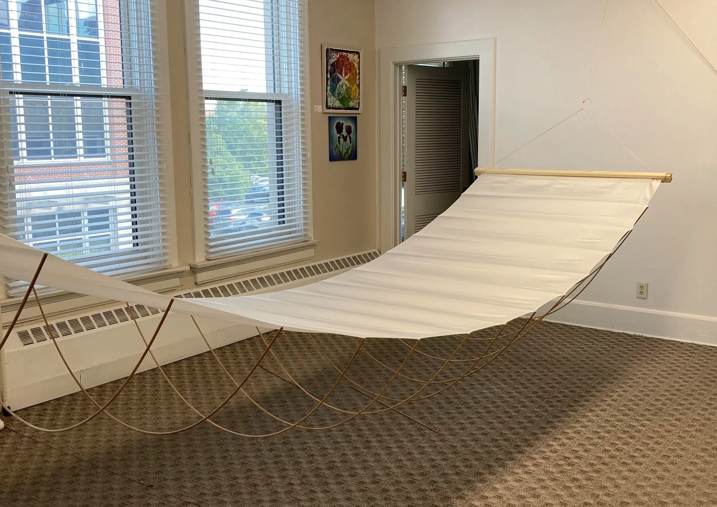 An art installation featuring a solitary white platform with a large, curved structure made of wooden sticks and string. The display is set in a room with large windows with blinds, artwork on the wall, and a doorway leading to another space. The room has a dark brown carpeted floor and off-white walls.