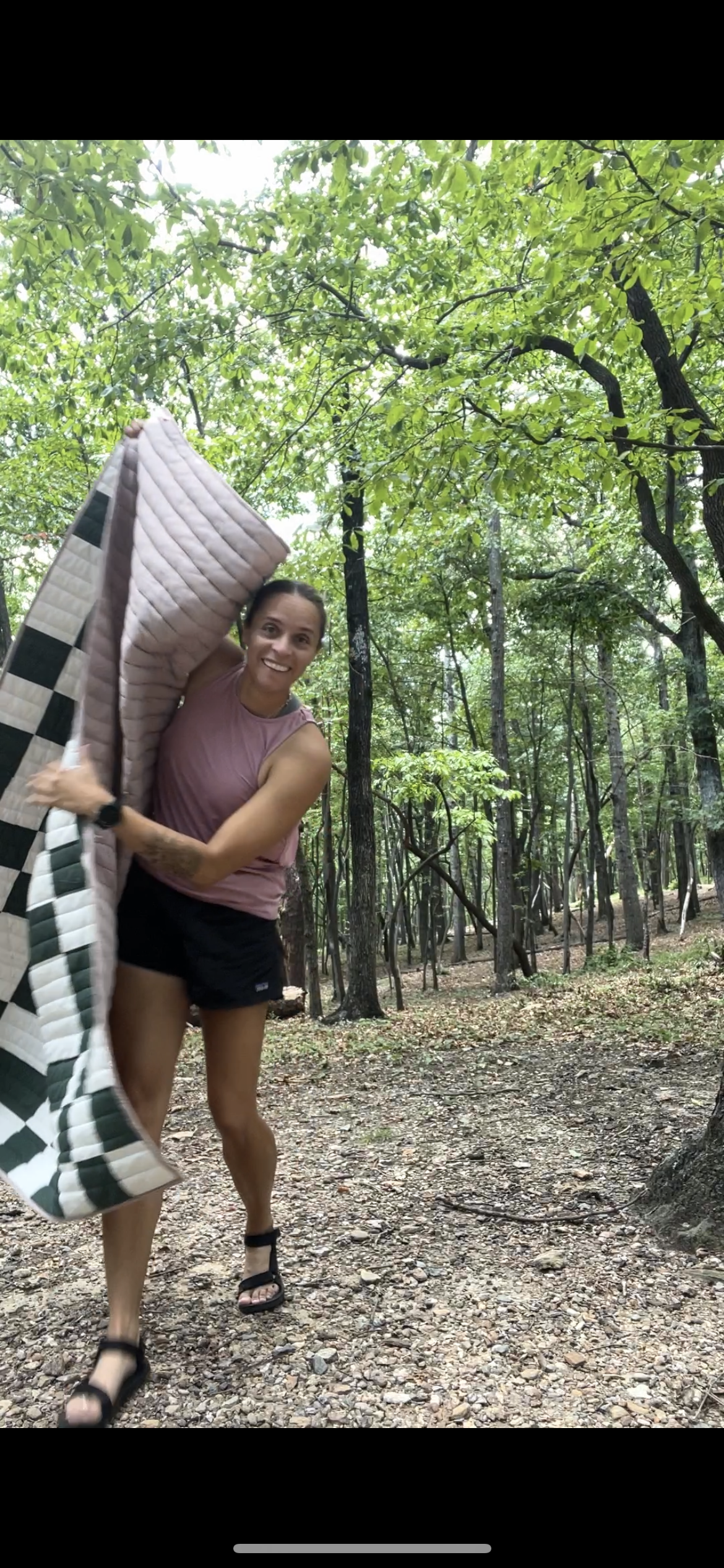 Woman in sleeveless pink top and black shorts holding a quilted blanket in a wooded park.
