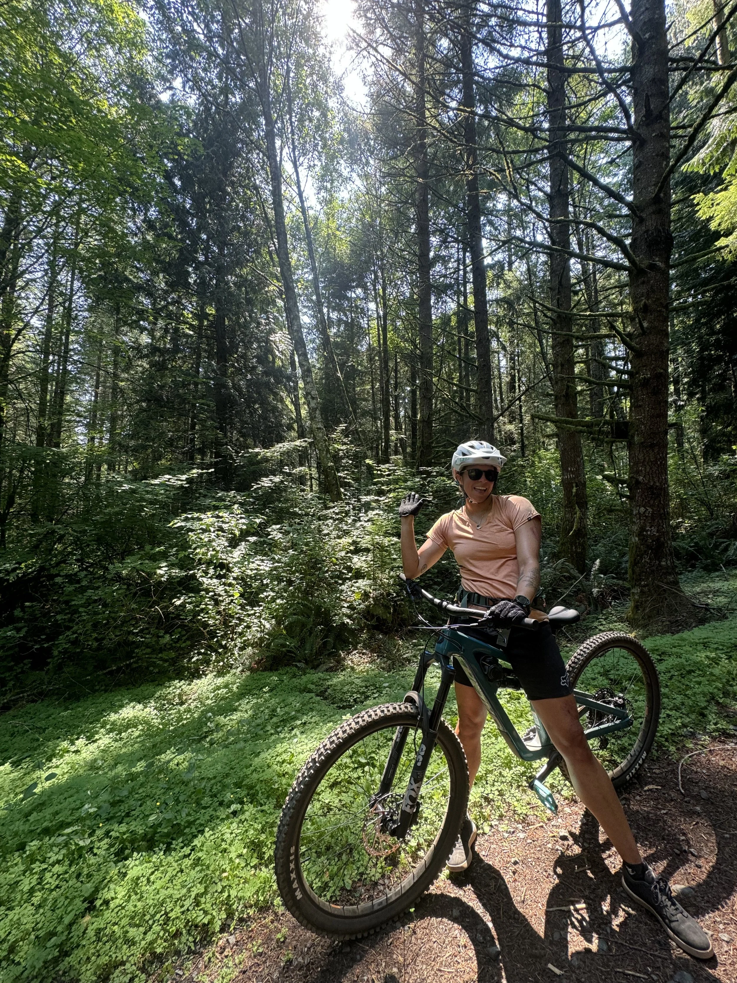 A woman standing with a mountain bike in a forest, wearing a helmet, sunglasses, a peach-colored shirt, black shorts, and gloves, smiling and waving.