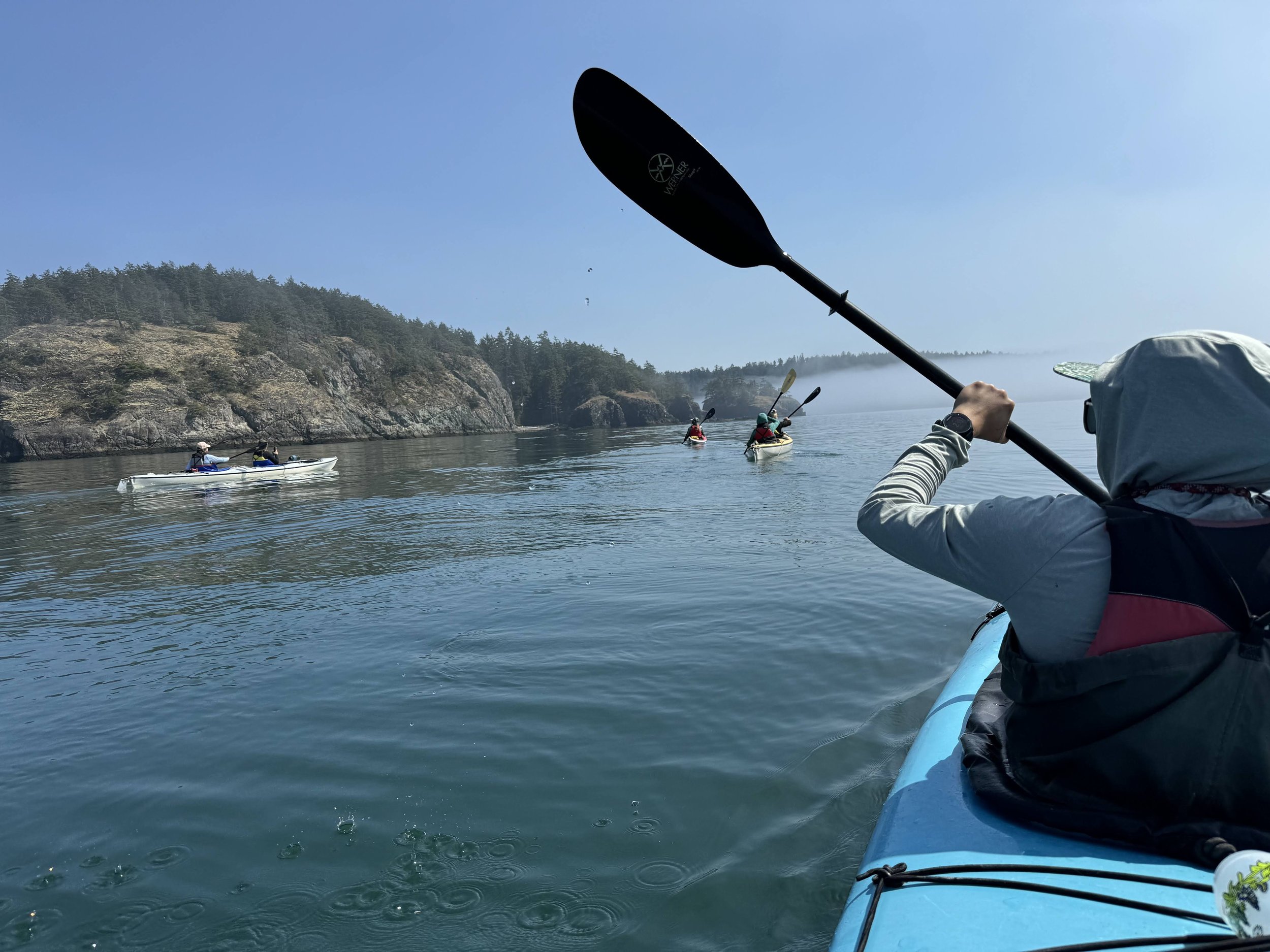 People kayaking on calm water near rocky and forested shoreline under a clear blue sky.