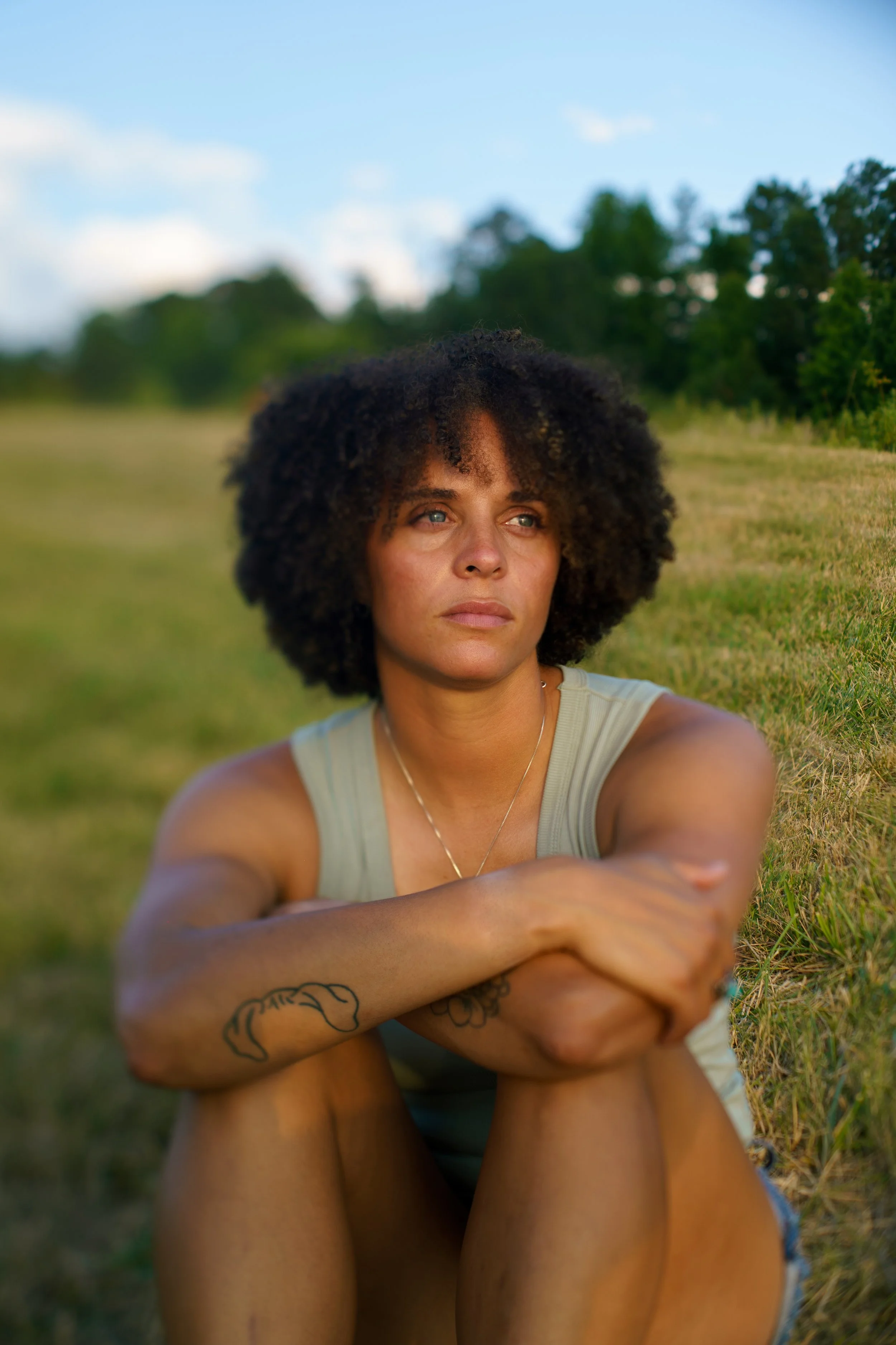A woman with curly dark hair sitting outdoors on grass, looking contemplative, wearing a sleeveless top, with trees in the background on a partly cloudy day.