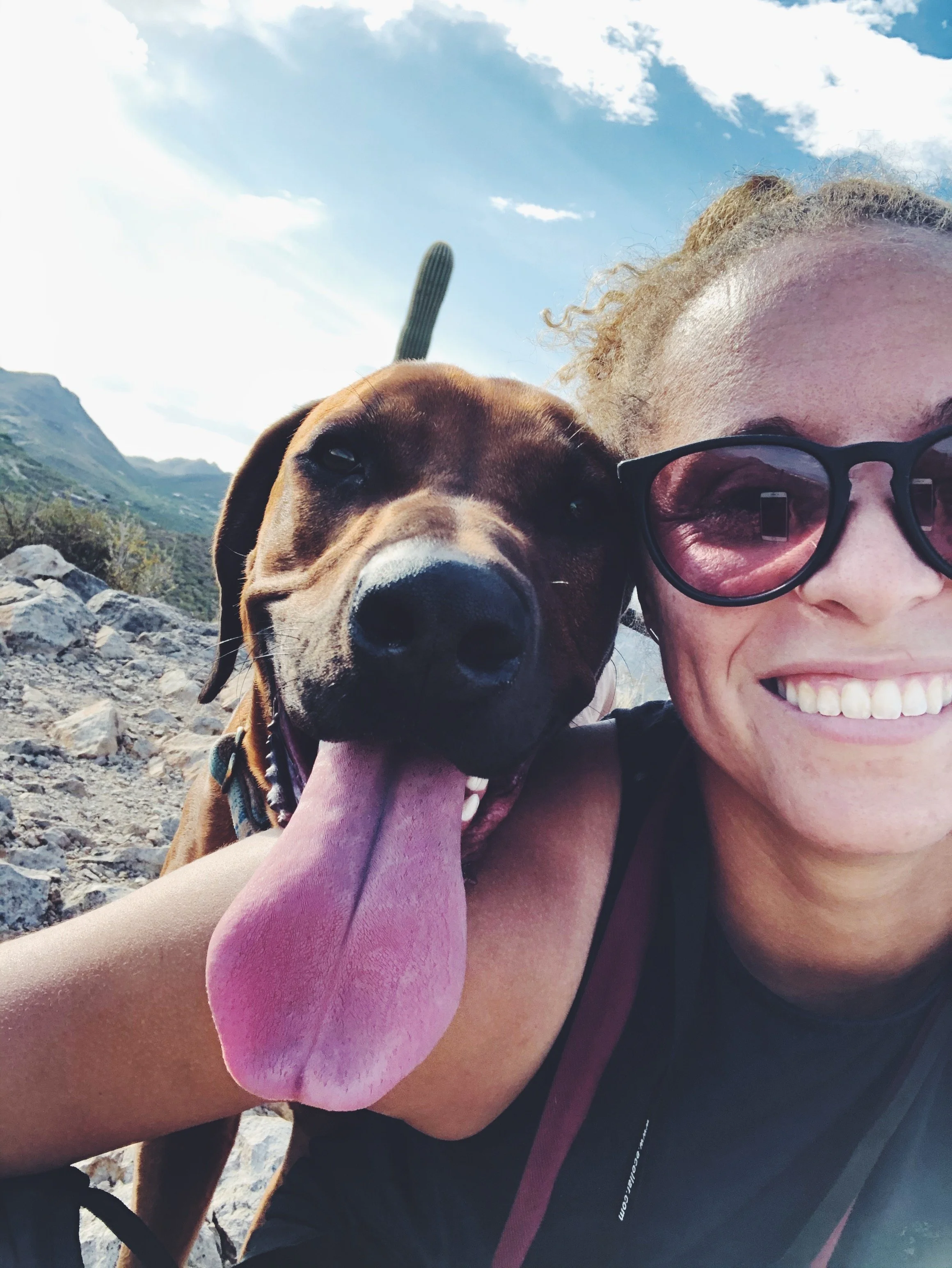 A woman smiling with sunglasses taking a selfie with her dog, a brown and black hound mix, outdoors with rocky terrain, mountains, and a blue sky with clouds in the background.