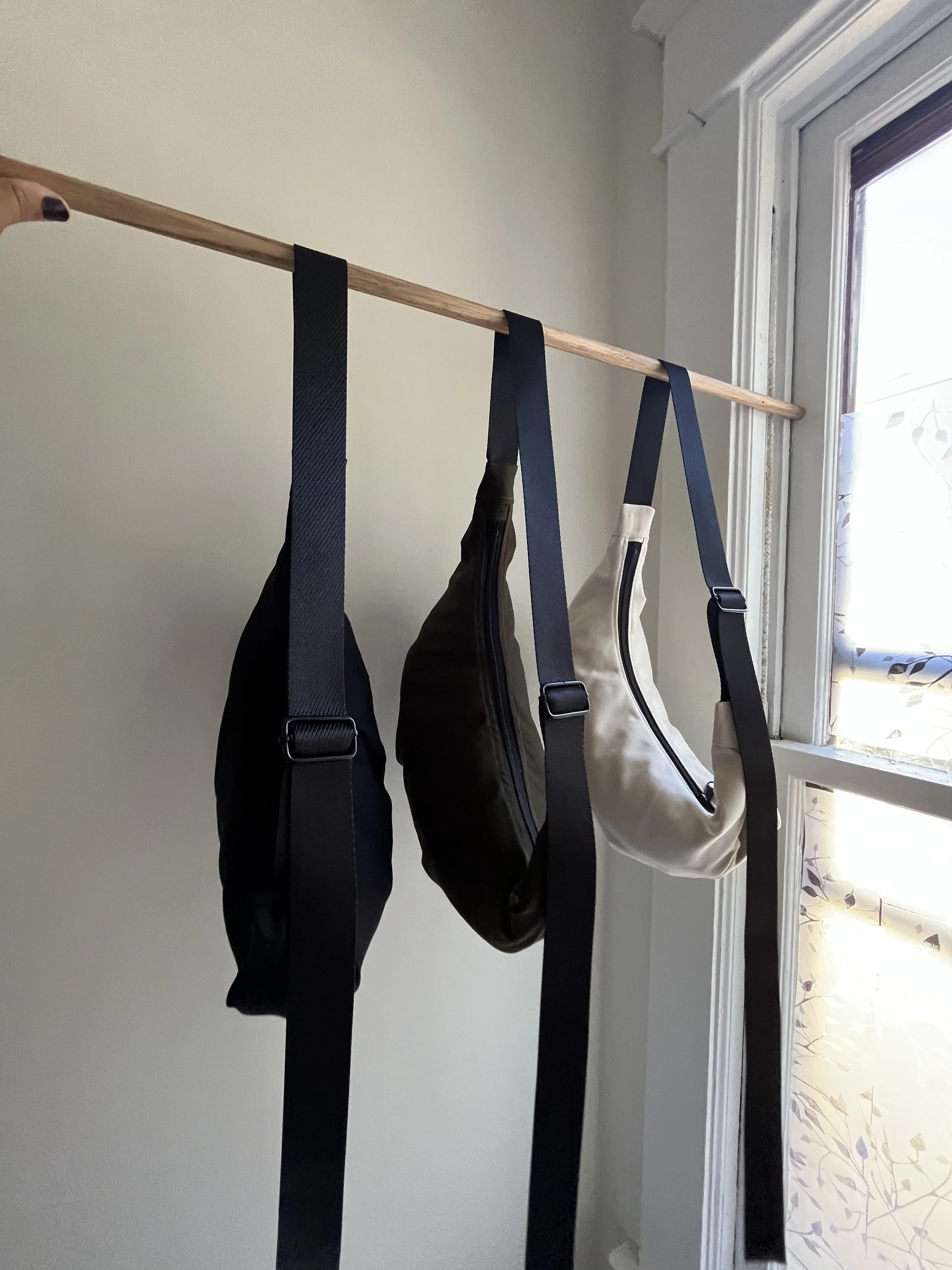 Three crossbody bags in black, brown, and beige hanging from a wooden rod near a window.
