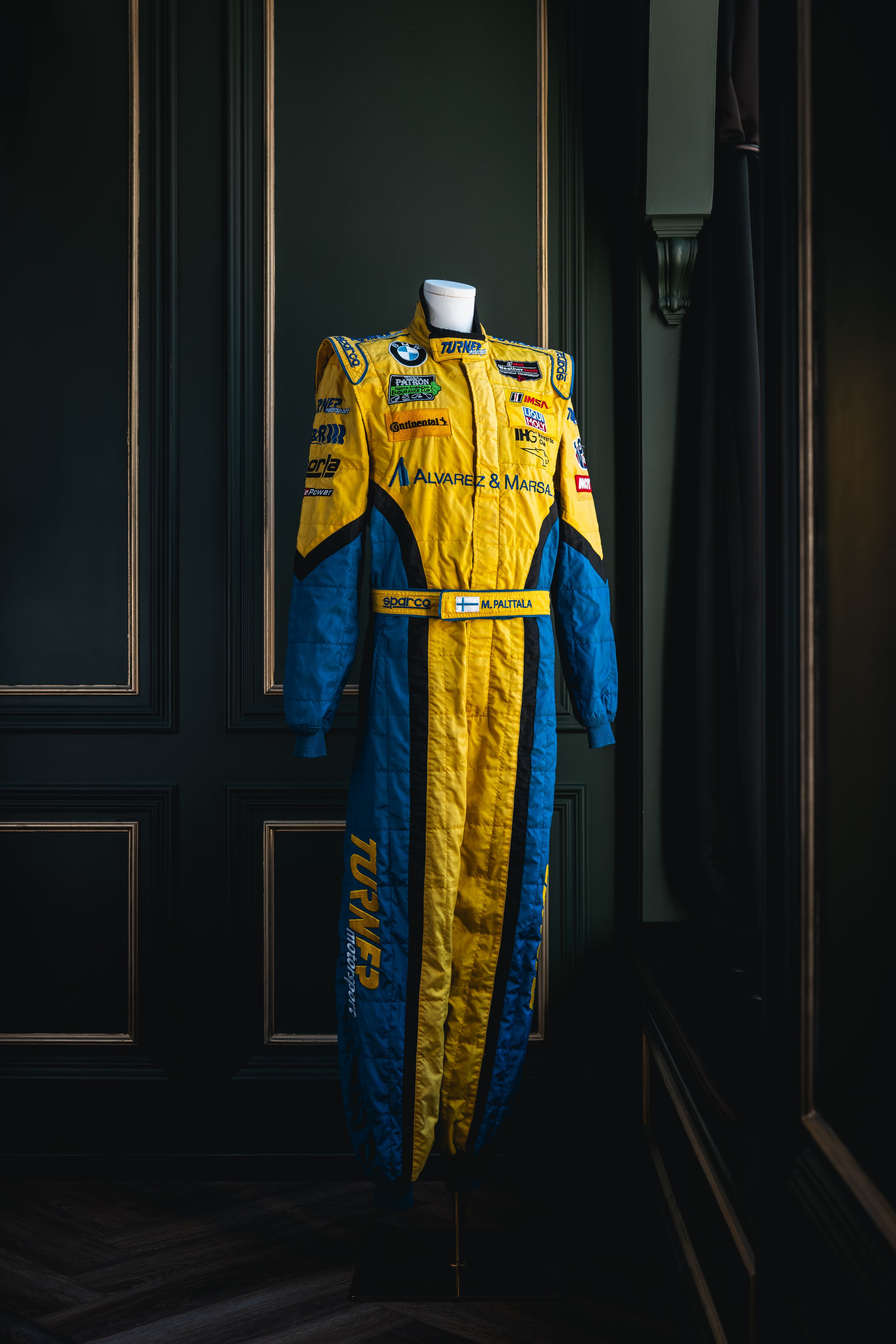 Race suit on display with yellow and blue colors, patches, and sponsors, featuring Finnish flags.