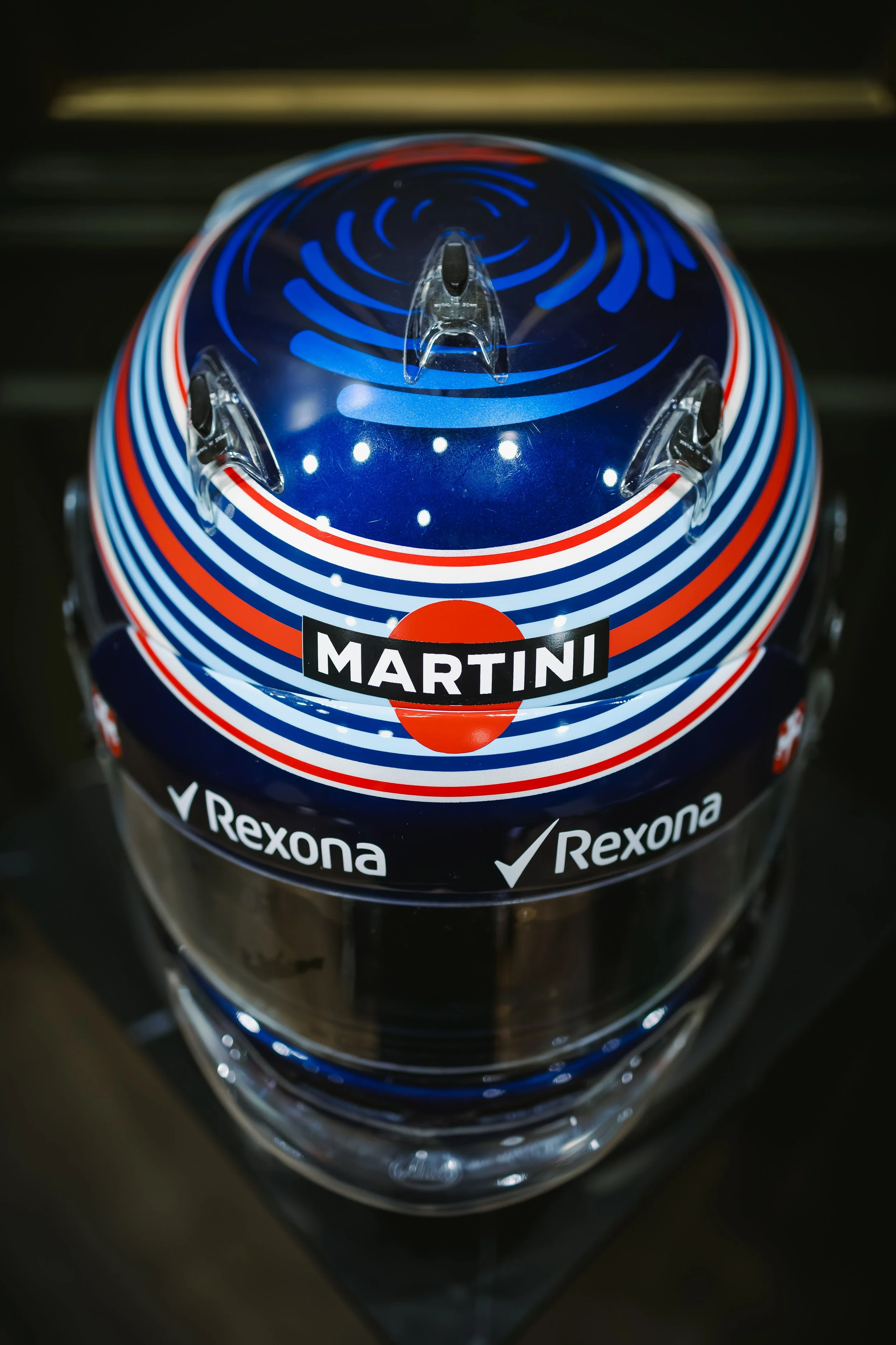 A race car driver helmet with a blue, white, and red striped design, featuring the Martini logo and Rexona sponsorship logos.