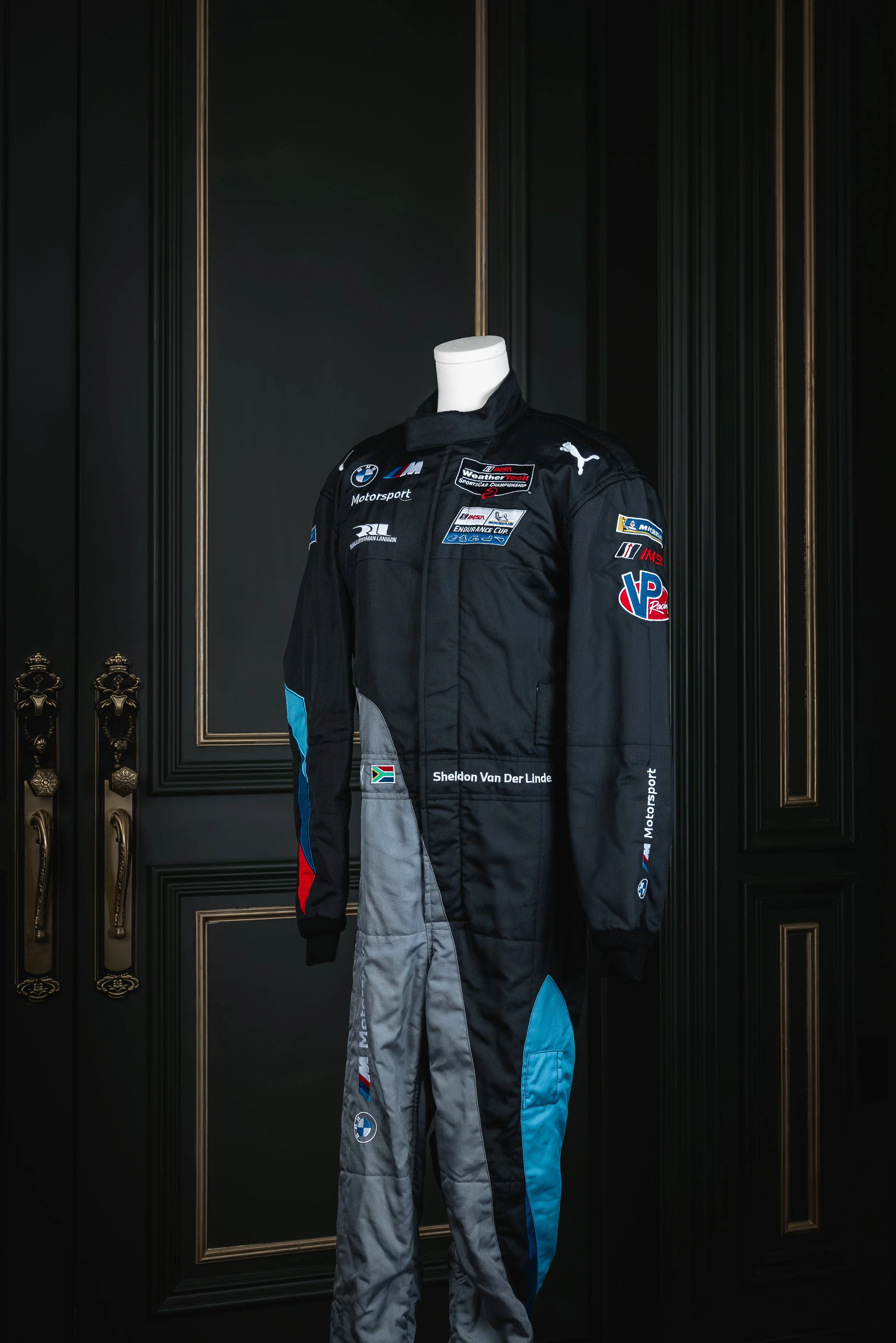 Race car driver in white racing suit with black accents, wearing a helmet, standing next to a blue race car in a pit lane.