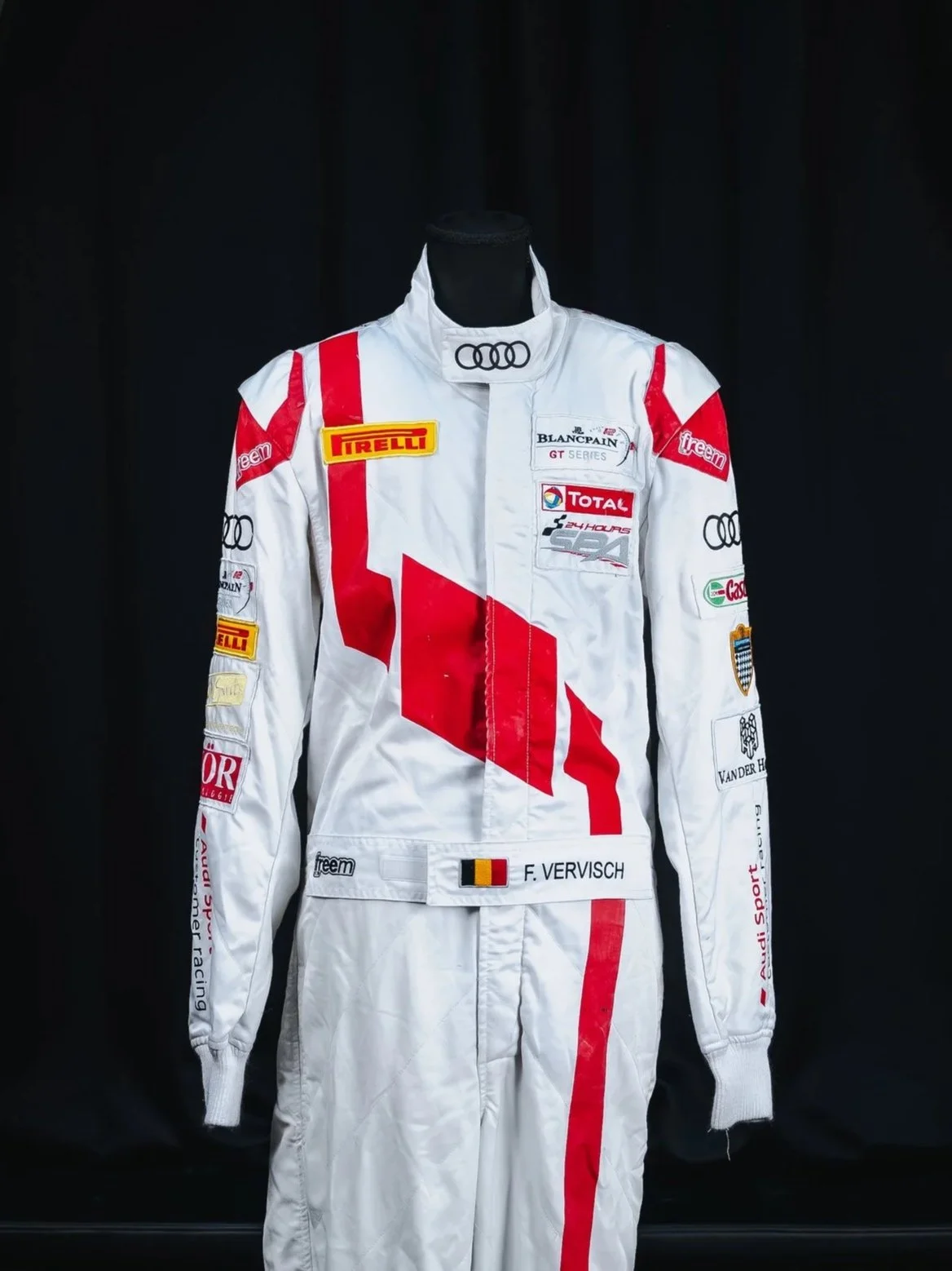 A white racing suit with red and black accents, displayed on a mannequin, with various sponsor patches and the name 'F. VERVISCH' on the belt.