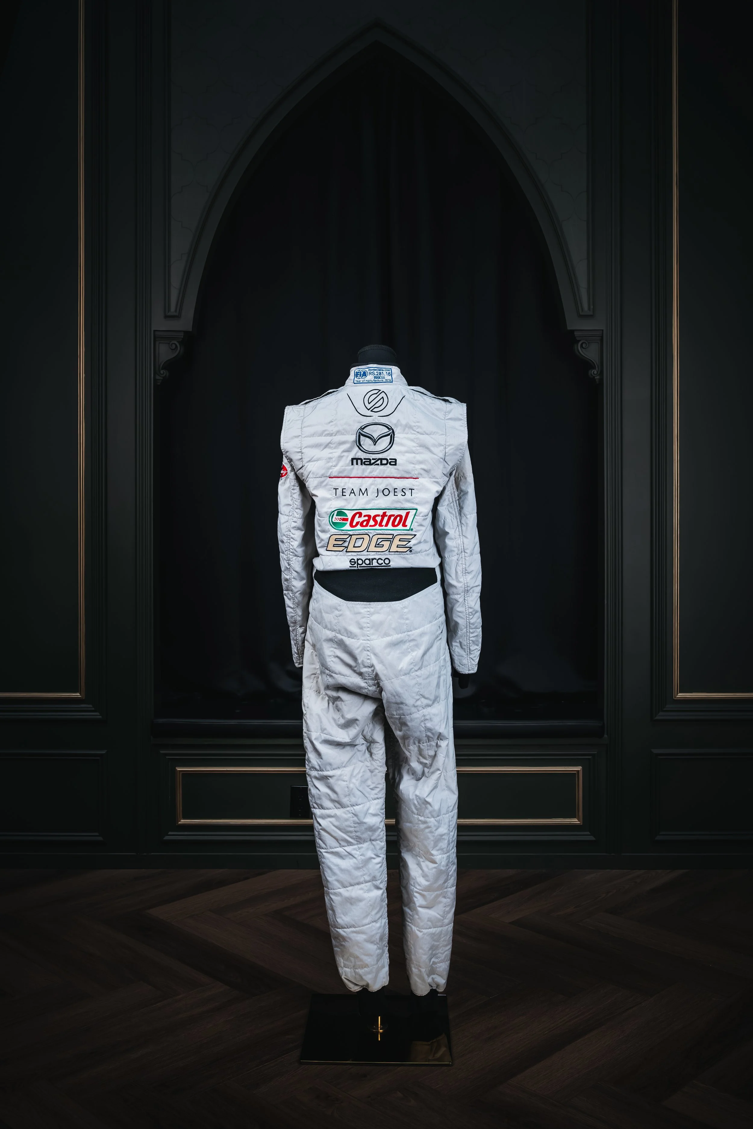 A racing suit on a mannequin display, facing away, with various sponsor logos including Mazda and Castrol, set against a black background with ornate frame details.