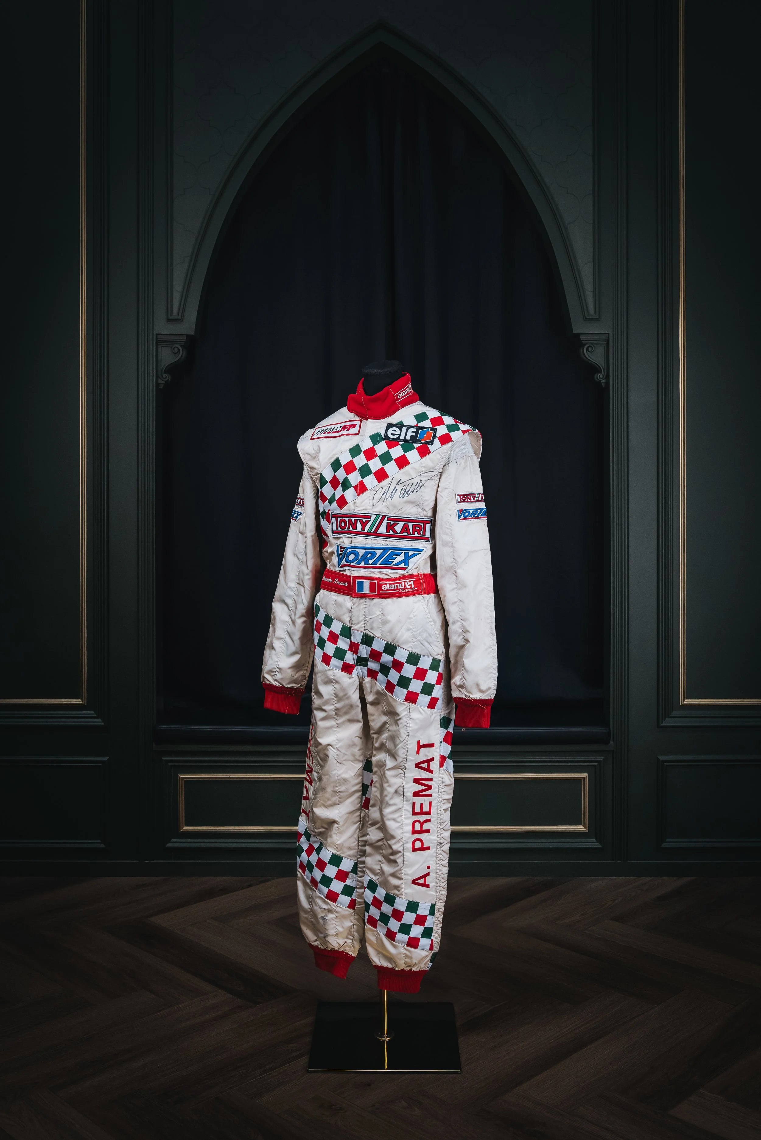 A vintage racing suit displayed on a mannequin in a dark, ornate room with gold accents. The suit is white with red, green, and blue logos and checkered patterns, signed on the chest.