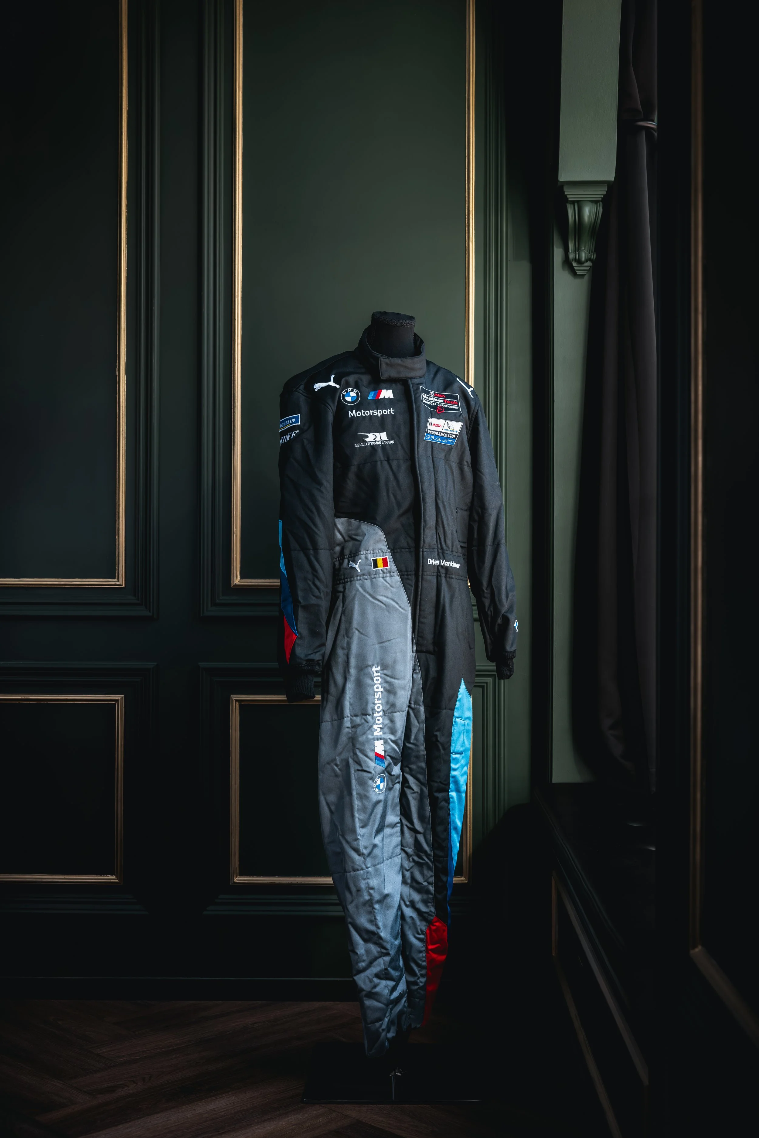 A racing suit displayed on a headless mannequin against dark green walls with ornate gold trim, near a window with dark curtains.