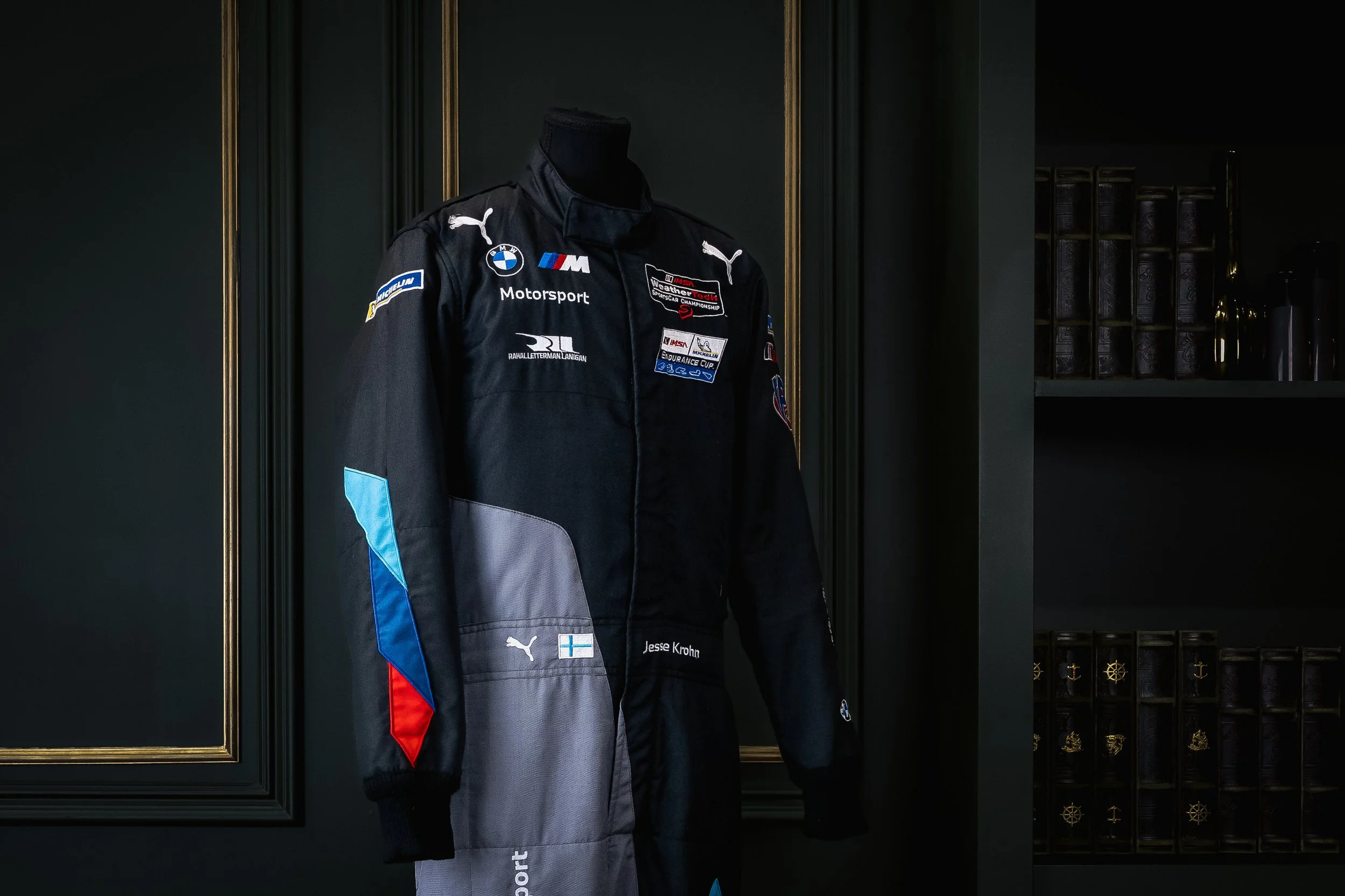 A black race car driver's racing suit on display, featuring various sponsor logos including BMW, Puma, and Michelin, with a Finnish flag patch and the name Jesse Krohn, in a dimly lit room with dark walls and gold decorative molding.