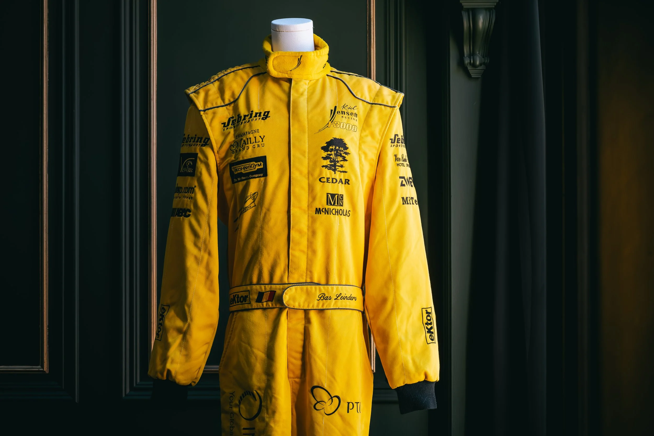 Yellow racing suit with various sponsor logos displayed on a mannequin against a dark background with wood paneling.