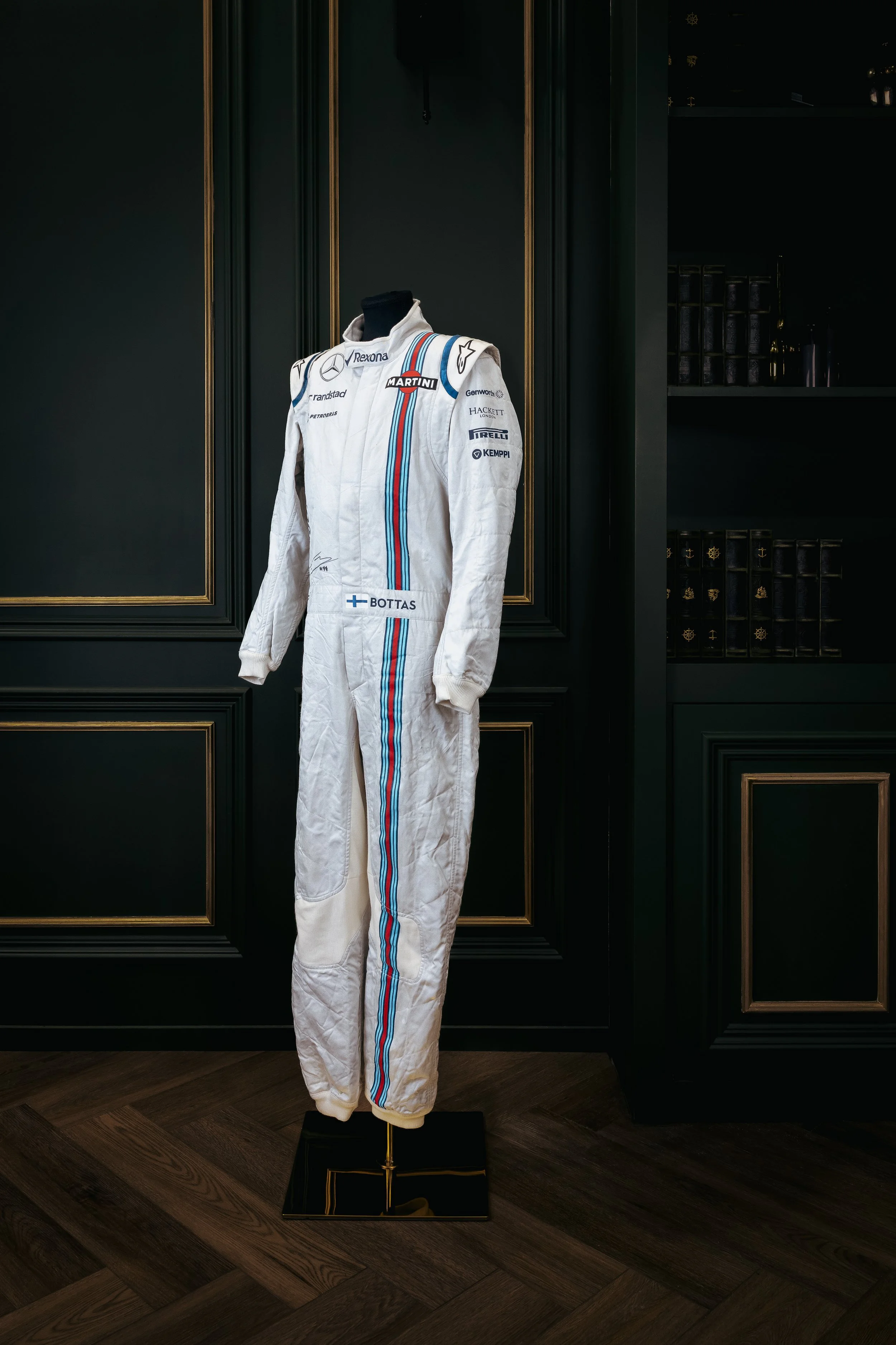 A white racing suit on display, inscribed with sponsor logos and the name Bottas, on a stand against a dark, ornate wall.