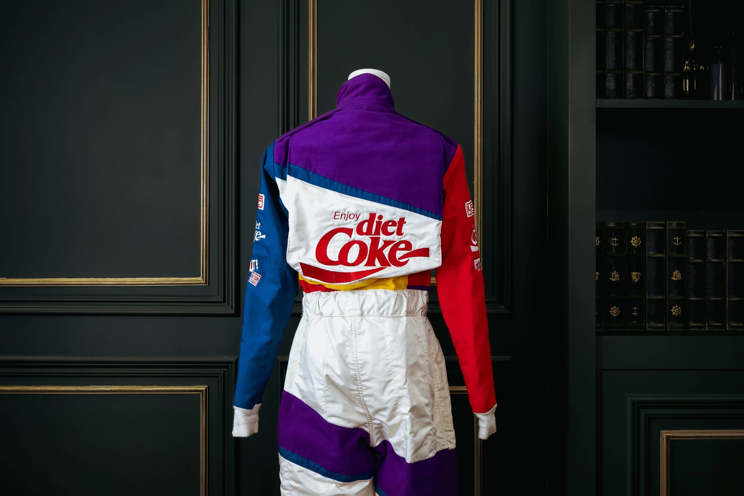 Colorful vintage racing suit on display with a "Diet Coke" logo on the back, set against a dark wall with gold accents.