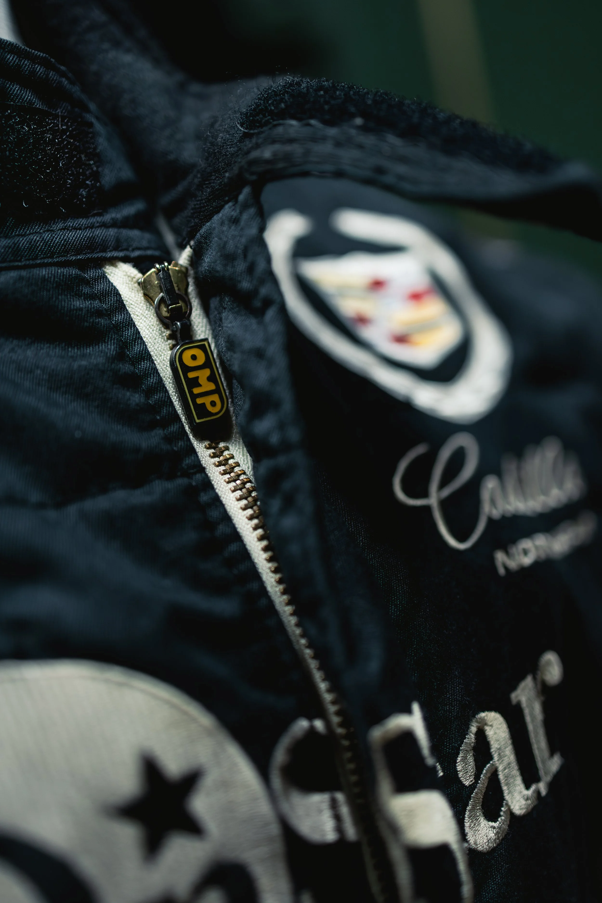 Close-up of a black jacket with a zipper that has a yellow and black OMP tag. The jacket features patches, including a circular patch with stars and other embroidered designs.