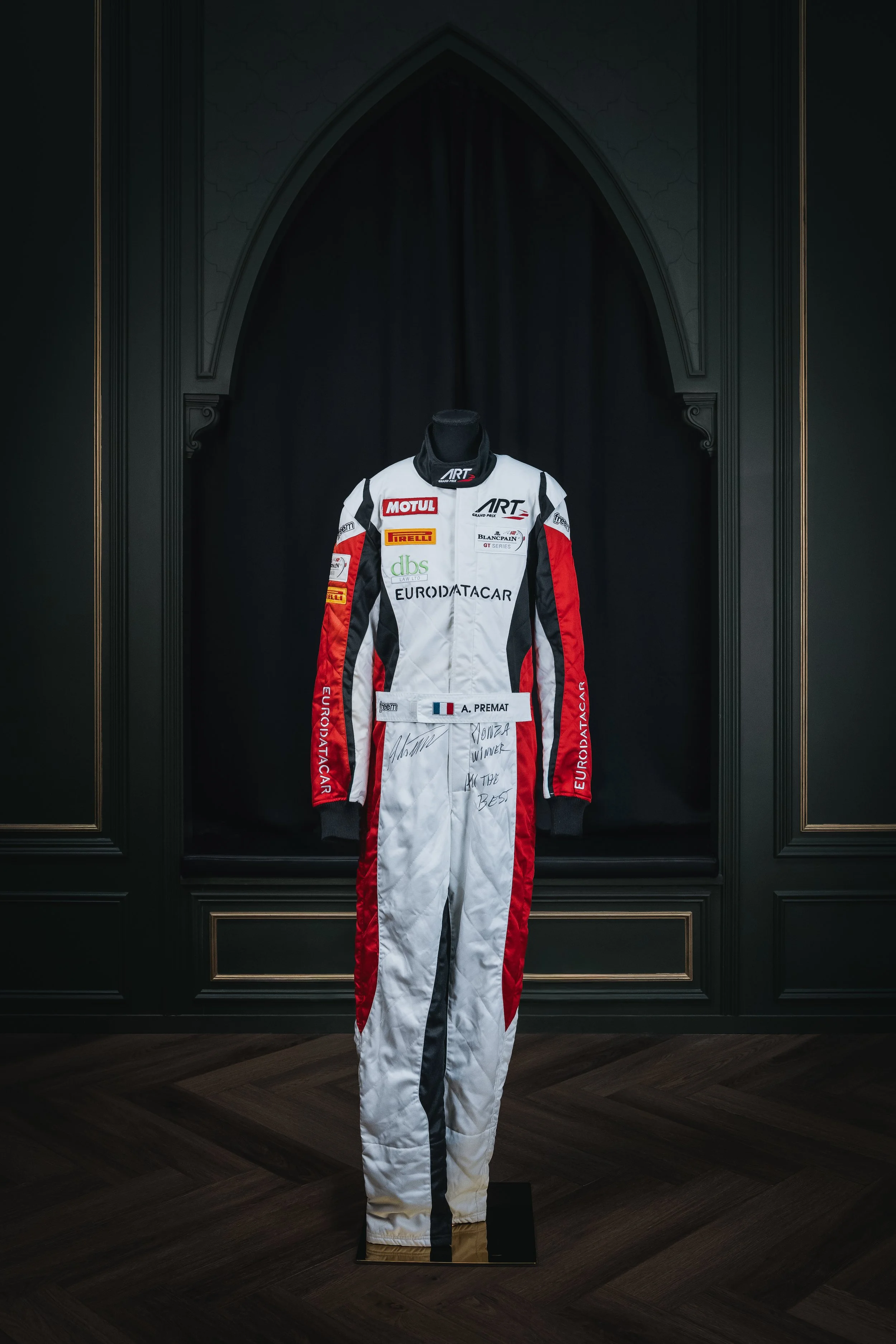 A racing suit displayed on a mannequin in a black frame with gold accents, on dark hardwood floor.