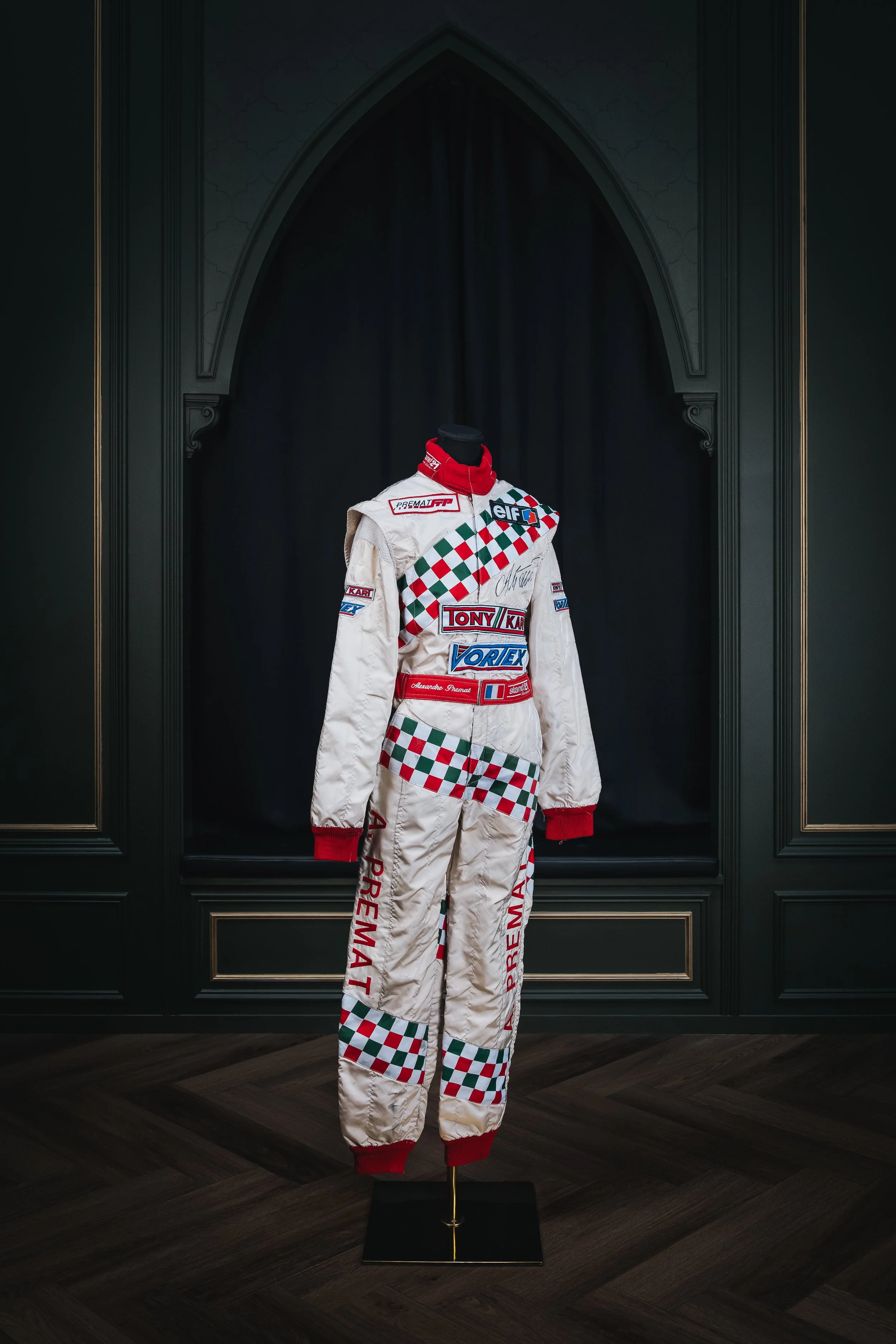 Racing suit displayed on a mannequin in front of a dark curtain, with a fancy black and gold ornate background.