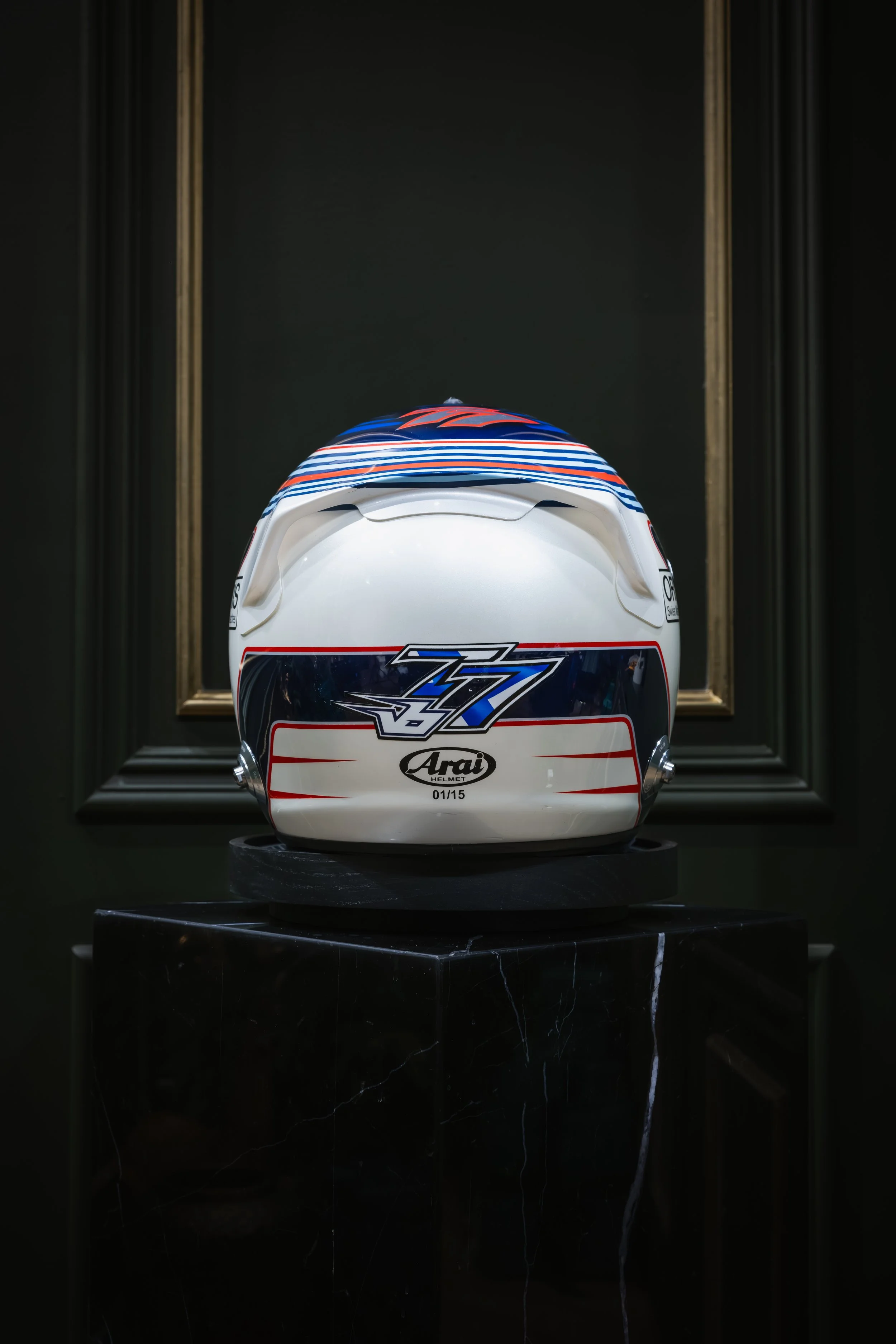 A racing helmet with the number 77 and the Arai logo, displayed on a black pedestal against a dark background.