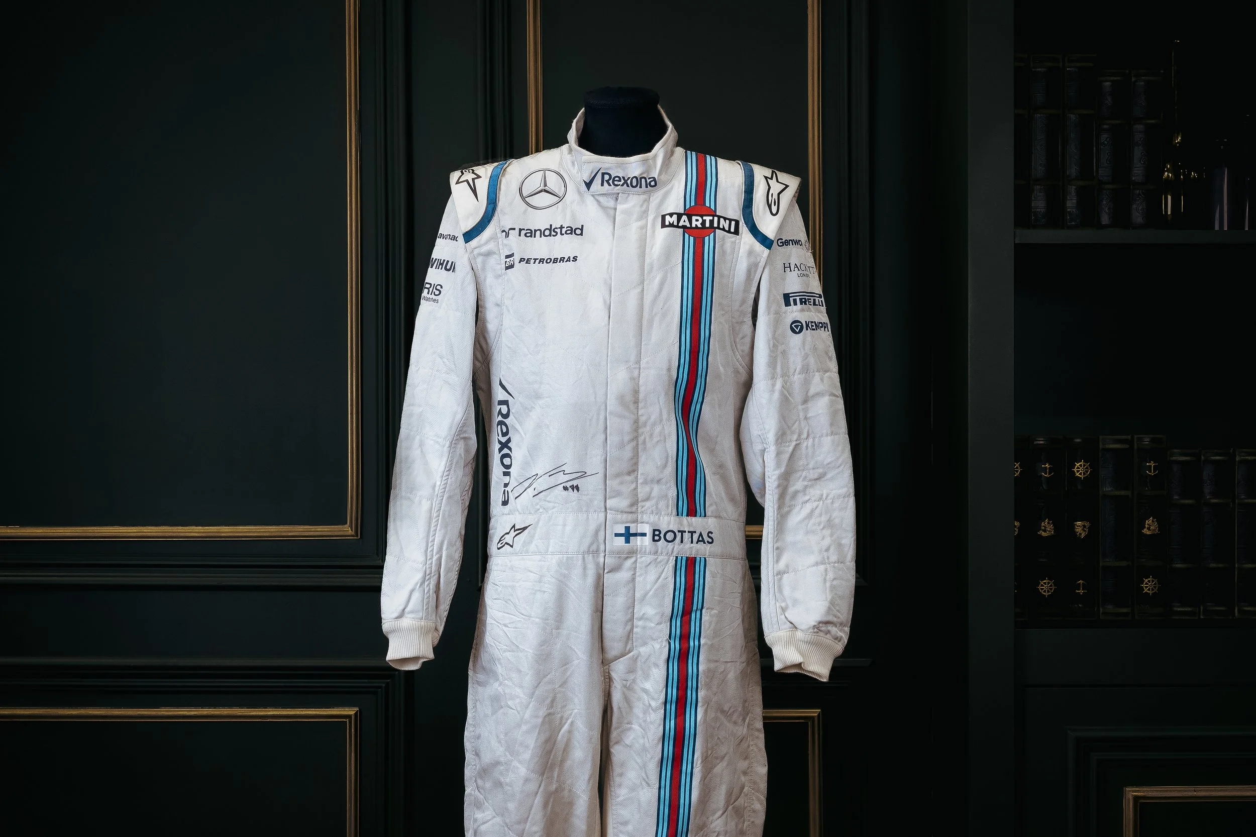 White racing suit with various sponsor logos and a multicolored stripe, displayed on a mannequin against a black wall with gold accents.