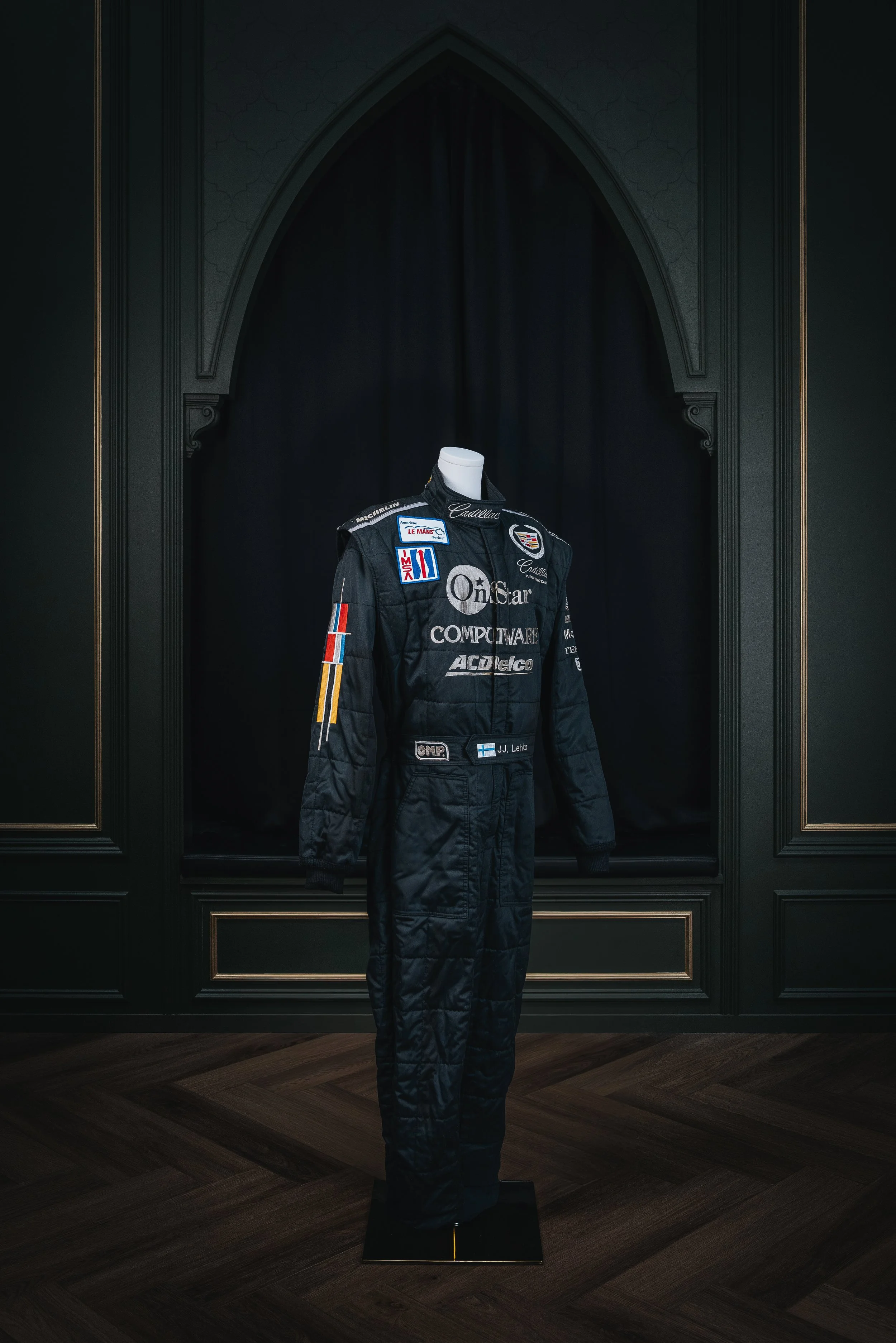 Racing driver's suit displayed on a mannequin in a dark room with ornate black and gold walls.