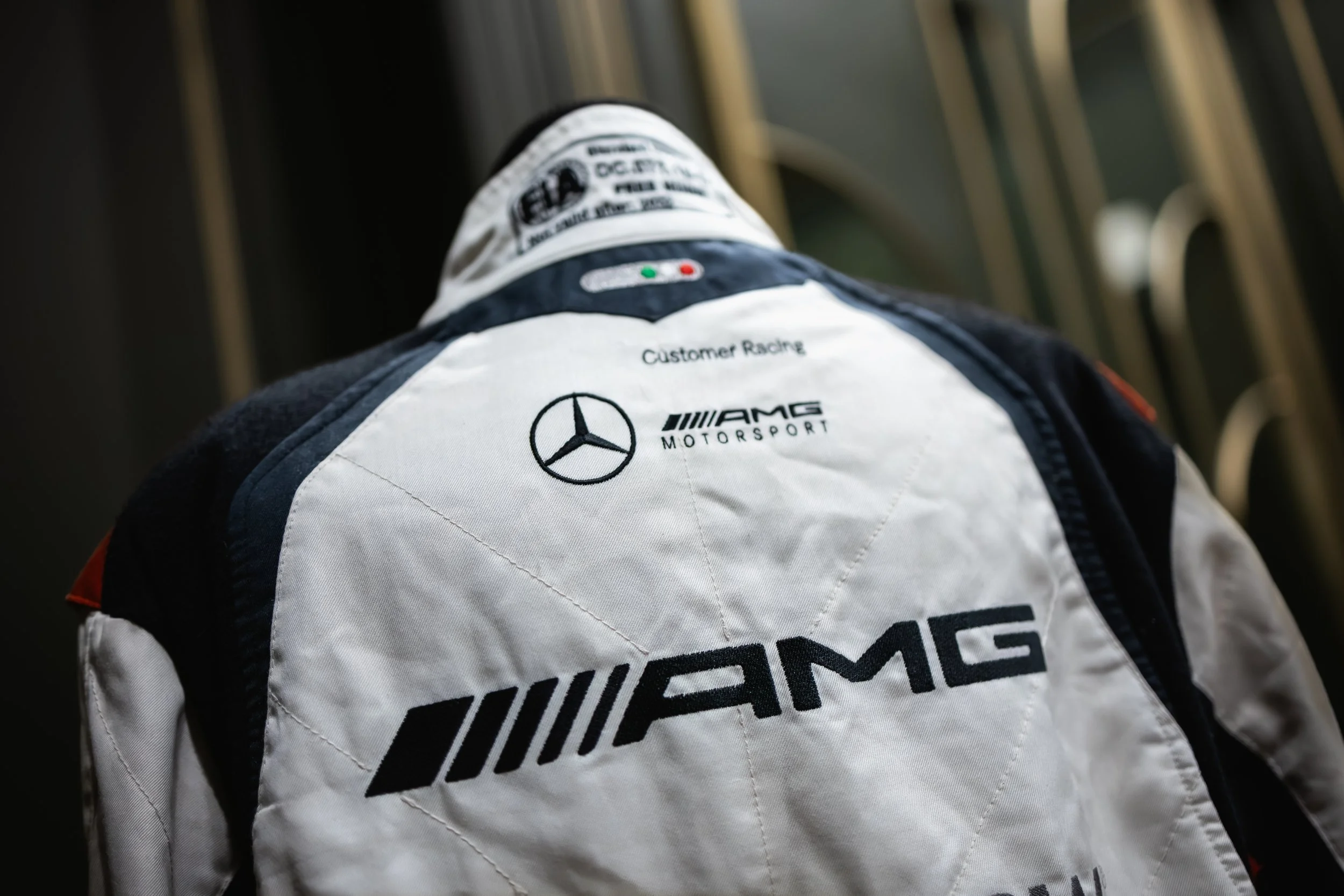 Close-up of a racing suit with Mercedes-Benz, AMG Motorsport, and customer racing logos, viewed from the back.