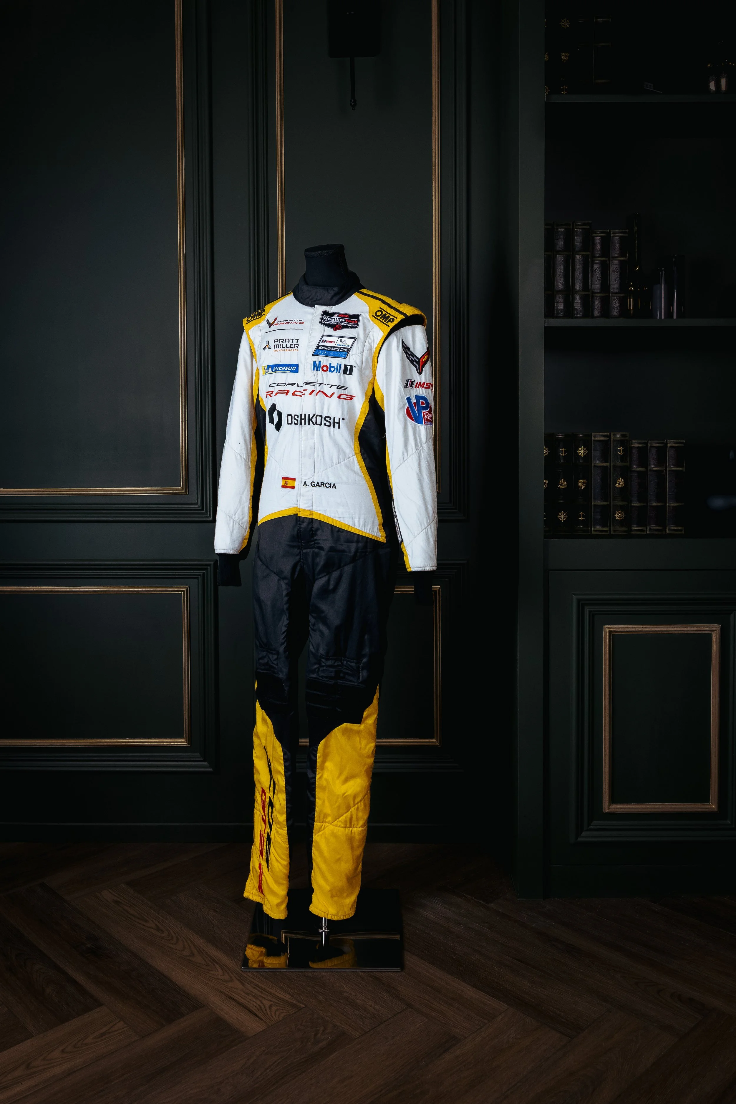 A racing suit displayed on a mannequin against a dark wall with wood paneling, featuring sponsor logos and a name patch reading "A. GARCIA".