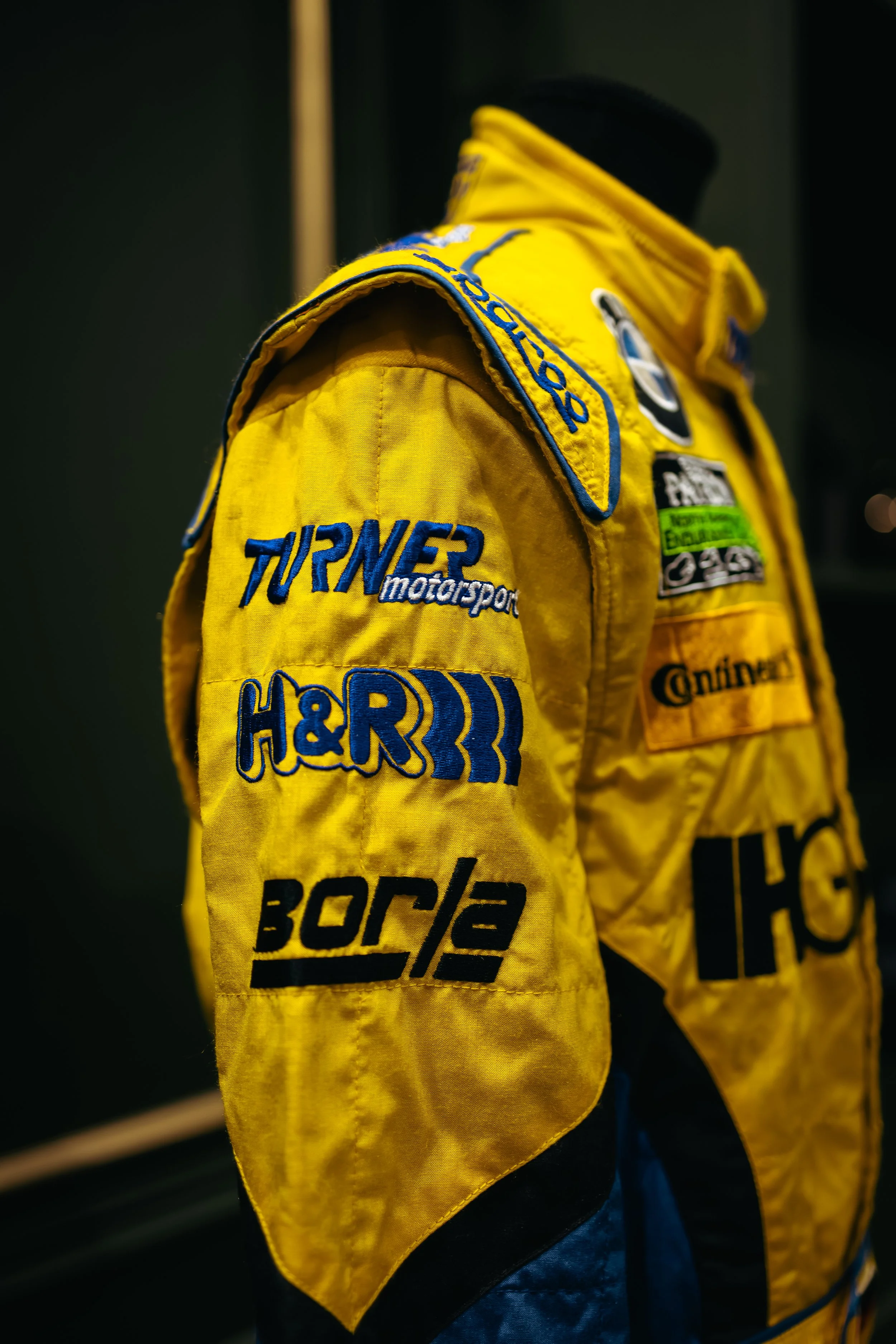 Close-up of a yellow racing suit with sponsor patches, including Turn 2 Motorsports, H&R, Continental, and Borla, displayed on a mannequin against a dark background.