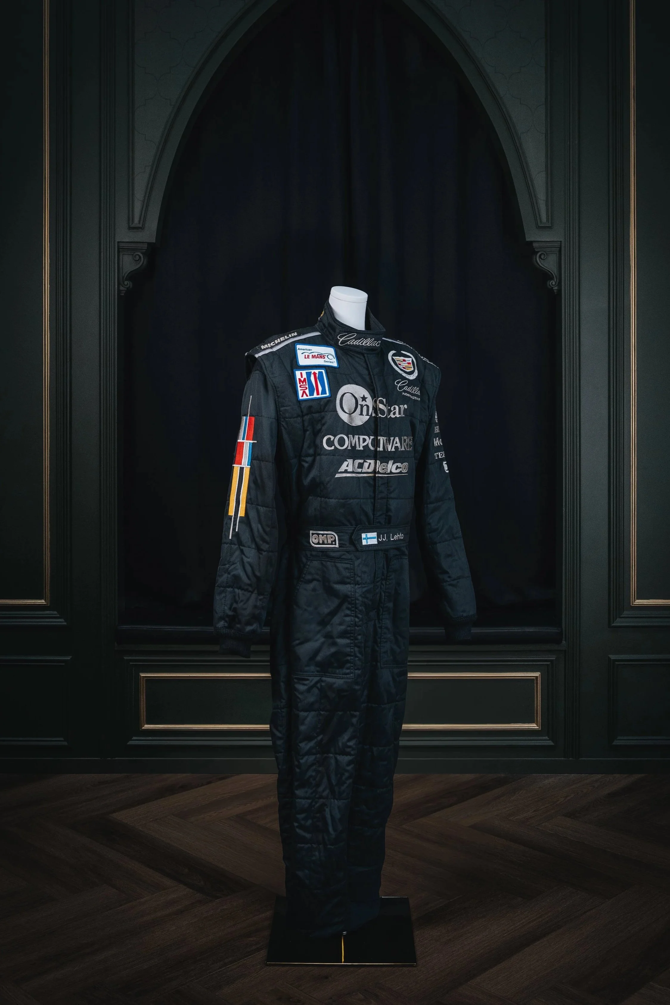 A black racing suit displayed on a mannequin in front of a dark backdrop, with various sponsor patches and logos, including Cadillac and Omsar, and a Finnish flag patch.