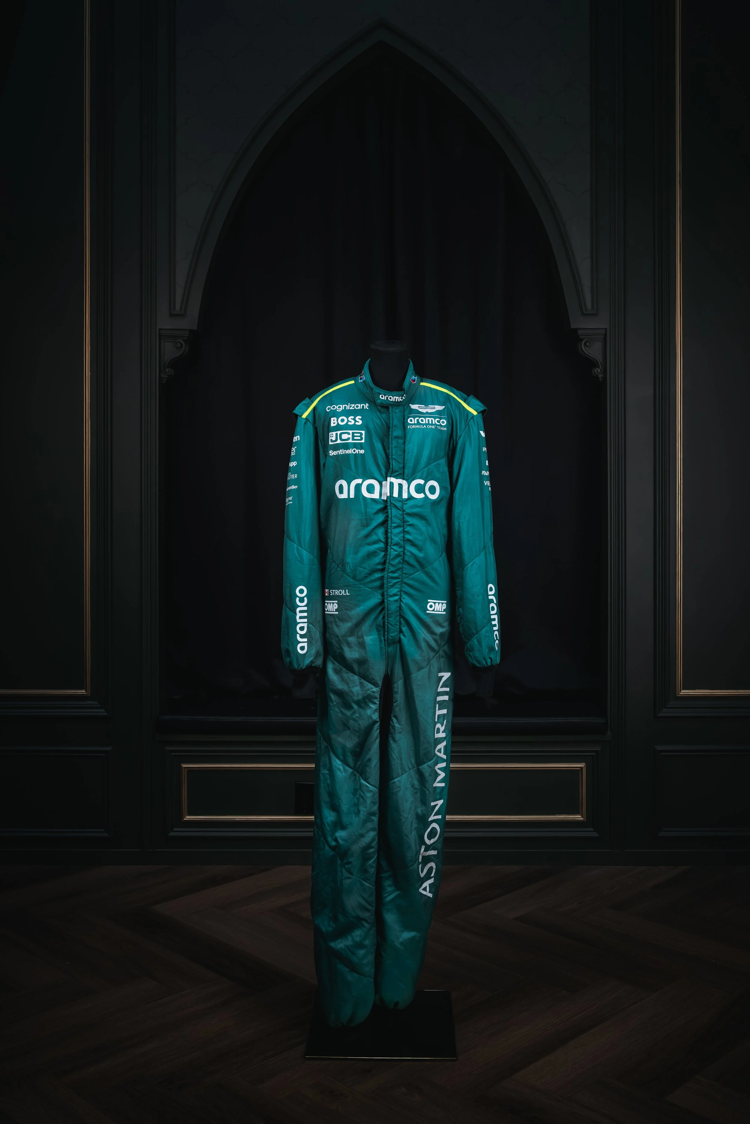 A racing suit displayed on a mannequin, featuring the Aston Martin logo and other sponsor logos, set against a dark background.