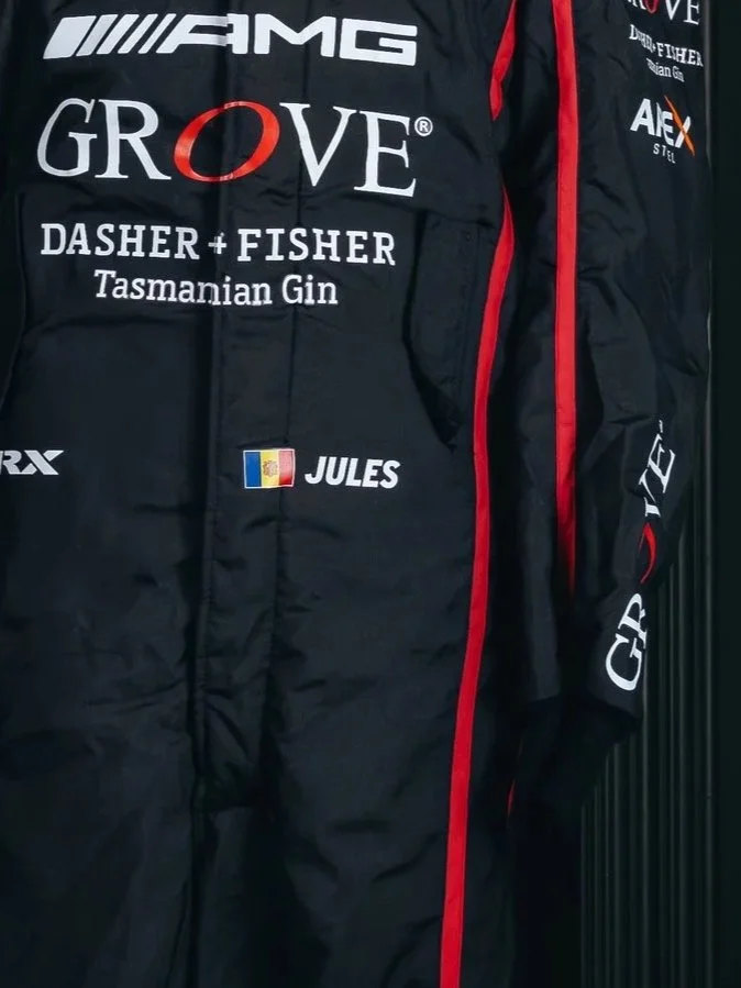 Close-up of a racing suit with various sponsor logos, including AMG, Grove, Dasher + Fisher Tasmanian Gin, Jules, and others. The suit is black with red accents and a French flag patch.