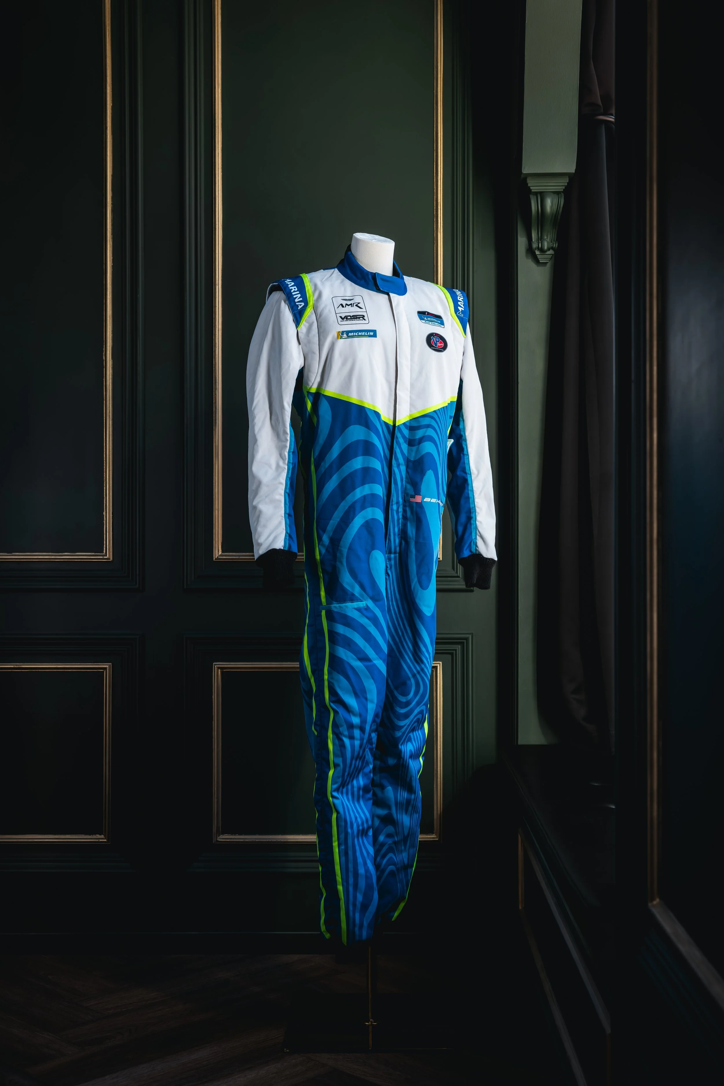 A racing jumpsuit on a mannequin, featuring blue, white, and yellow colors with logos, displayed in a room with dark green walls and wooden paneling.