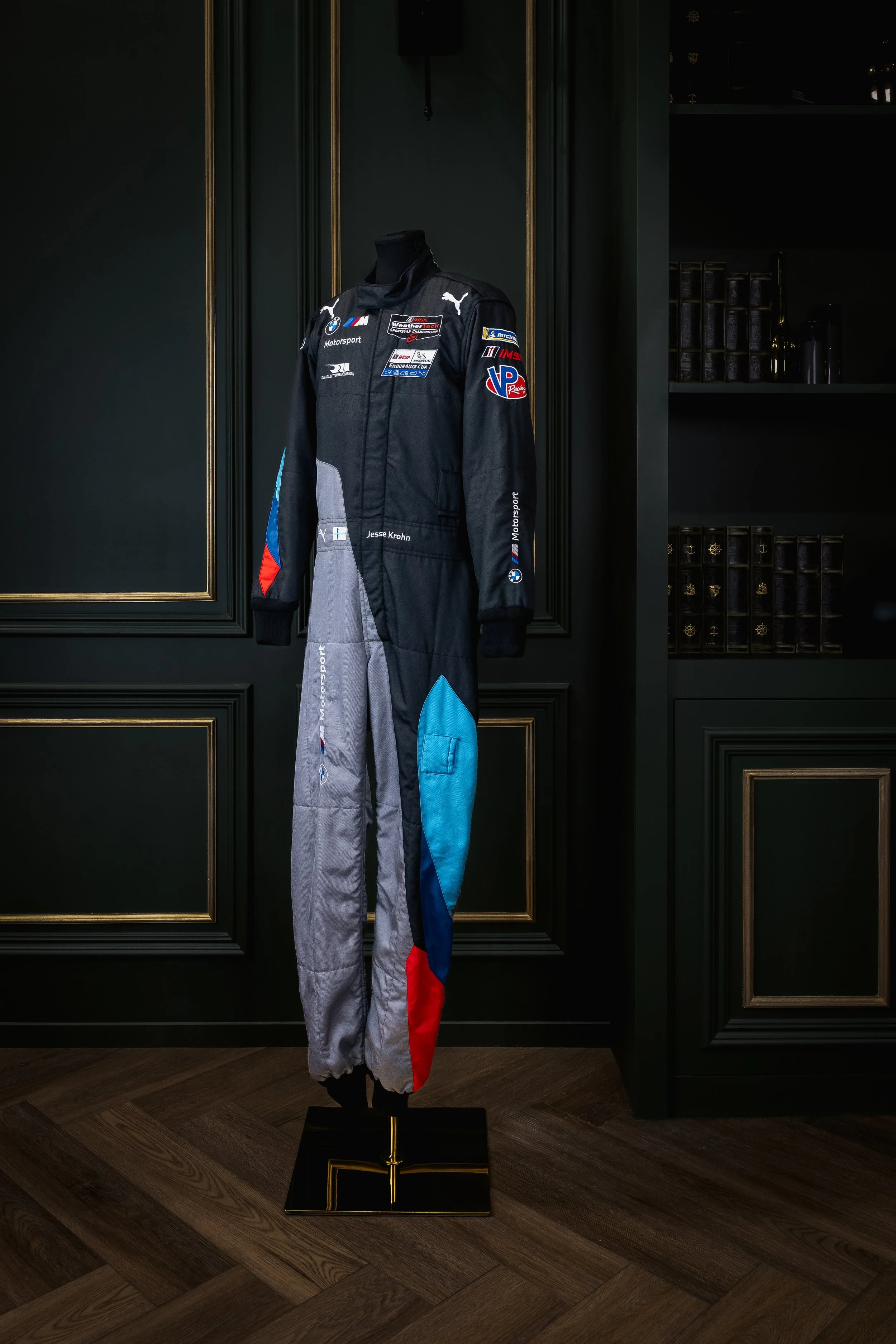 A racing driver's suit displayed on a mannequin, with patches including BMW, Puma, and other sponsors, as well as the name Jesse Krohn, against dark-paneled walls with gold trim in a room with wooden floors.